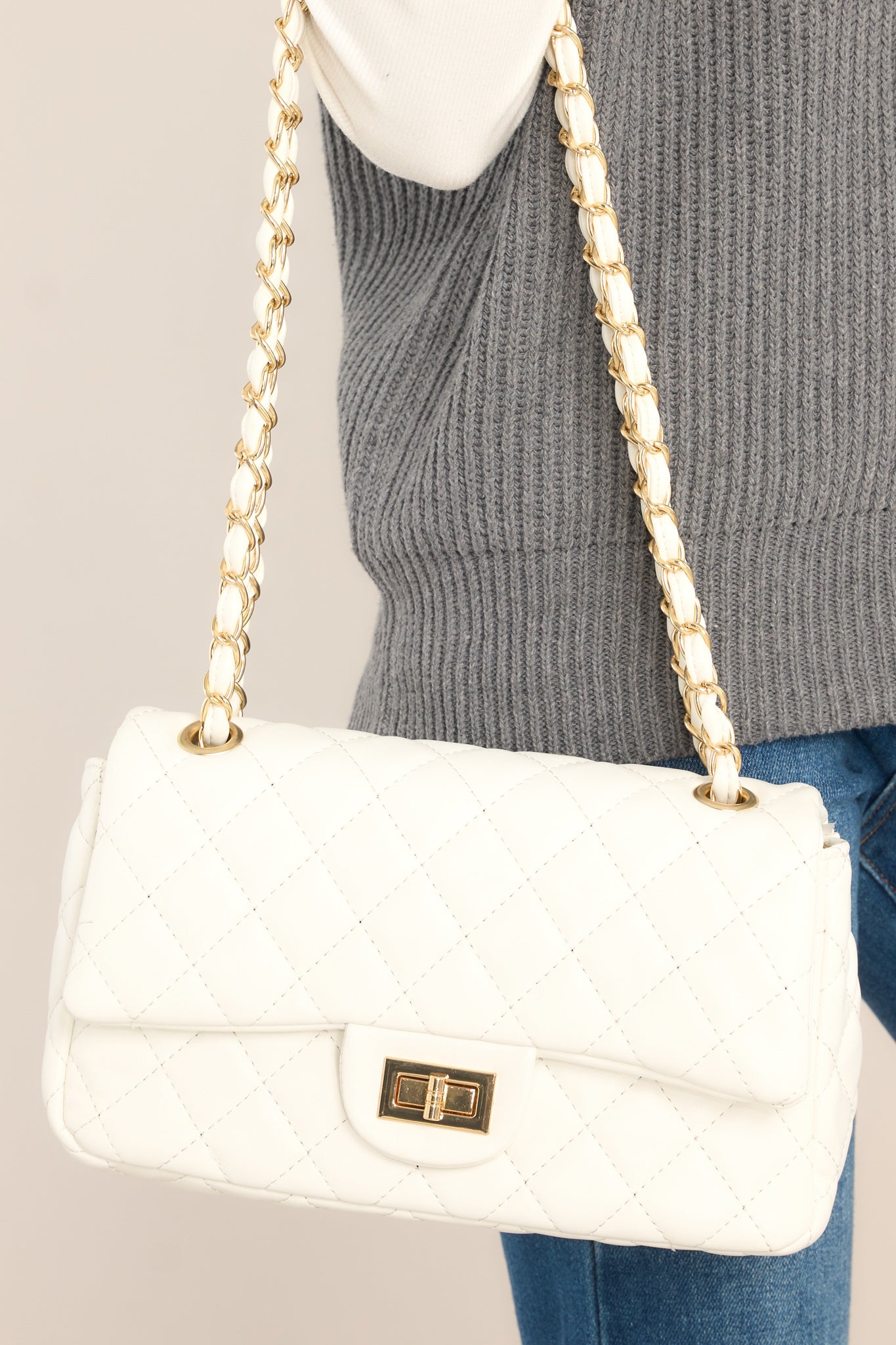 This white hang bag features a single flap, a chain strap, a turn lock closure, a hidden pocket, a zippered pocket on the inside, and gold hardware. 