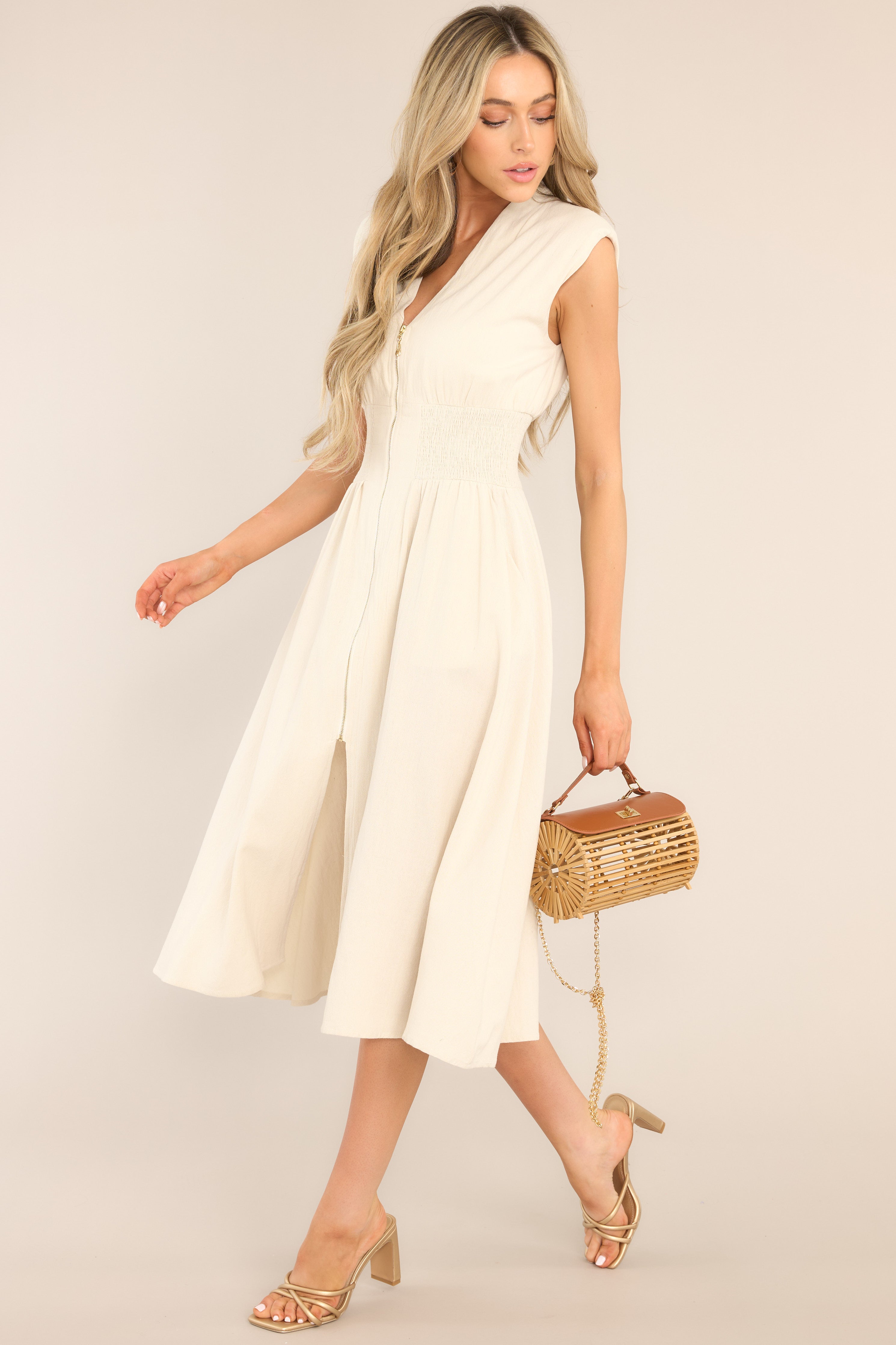 This beige dress features a deep v-neckline, shoulder padding, a functional zipper front, a fully smocked waist, functional pockets, and a front slit. 