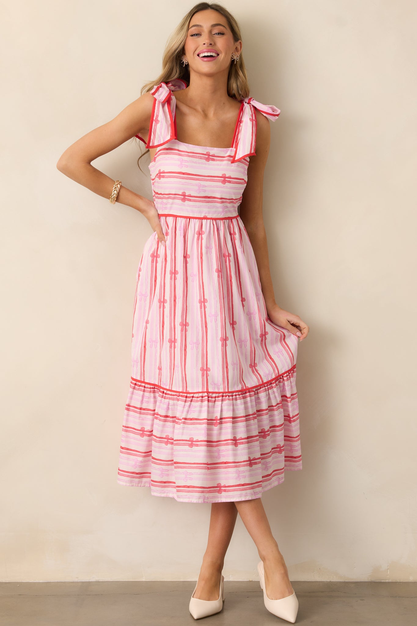 A full view of the pink dress featuring a square neckline, red trim, and self-tie shoulder straps, with a tiered bottom and bow stripe print.