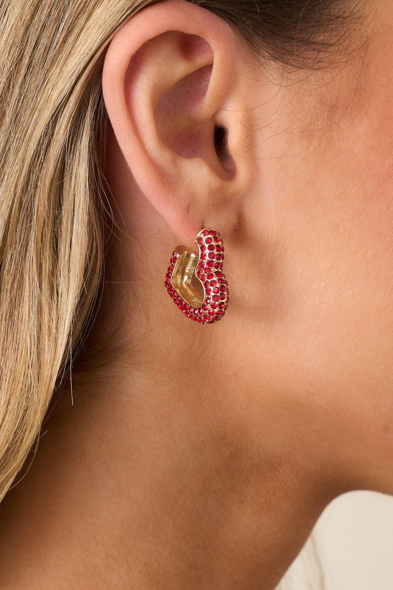 A detailed view of the pair of earrings, highlighting their incomplete hoop design, gold hardware, and secure back postings with shimmering red rhinestones.