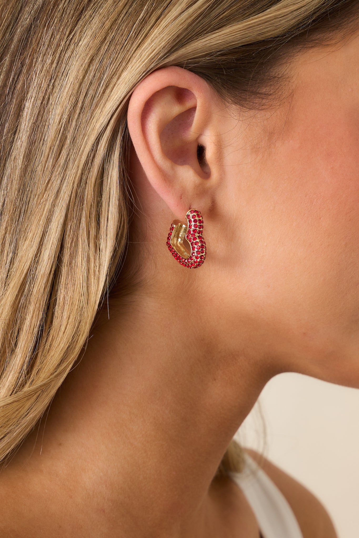 A close-up view of one red rhinestone earring, featuring a sideways heart shape, gold hardware, and small red rhinestones embedded in the design.