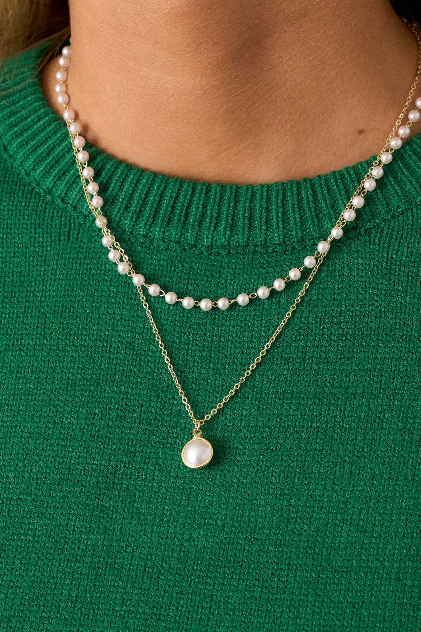 Gold layer necklace; the first layer is made of faux pearls strung together, while the second layer consists of a dainty gold chain holding a singular, larger faux pearl, all held together by a lobster claw clasp.