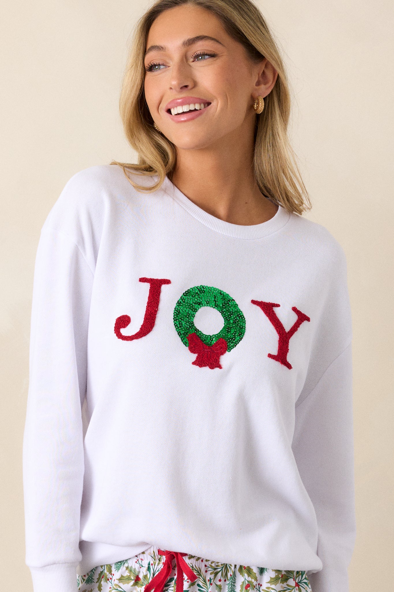 Tucked in view of a white sweatshirt featuring a crew neckline, Joy detailing with a wreath accent, a relaxed fit, and long sleeves with ribbed hems.