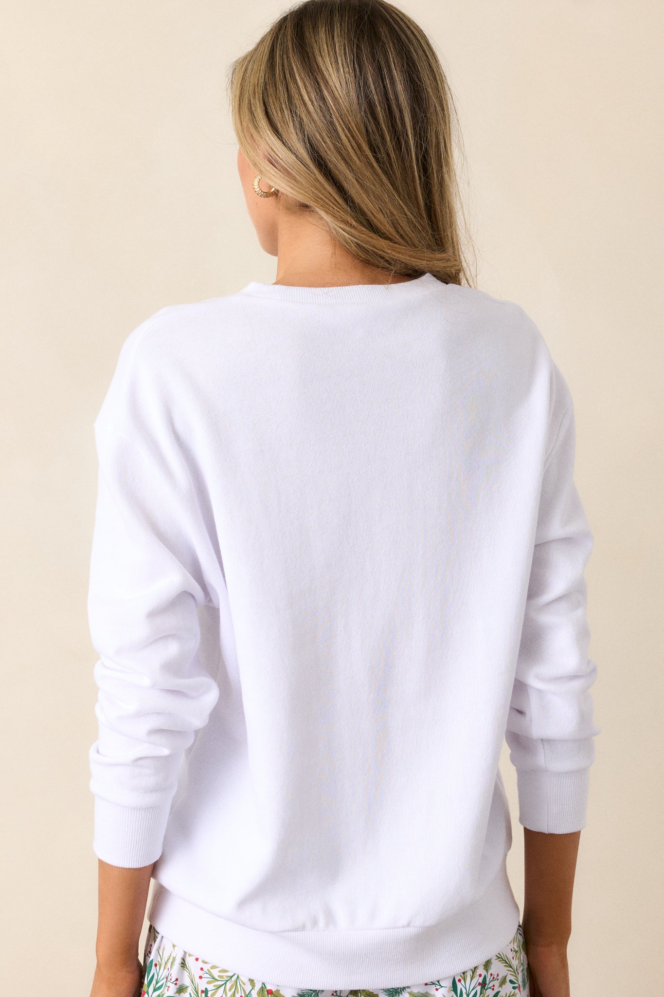 Back view of a white sweatshirt featuring a crew neckline, Joy detailing with a wreath accent, a relaxed fit, and long sleeves with ribbed hems.