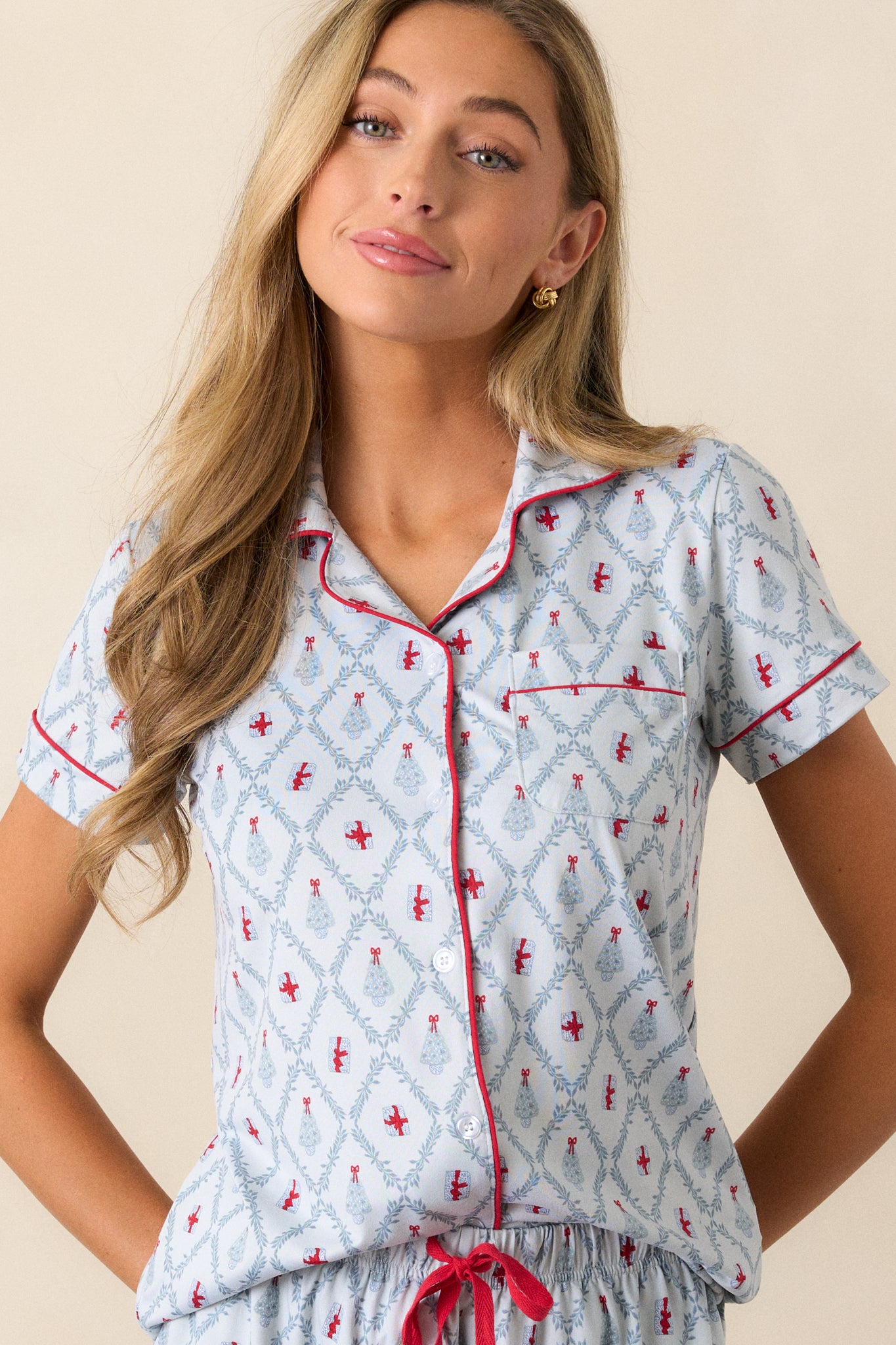 Front view of a light blue pajama shirt featuring a collared neckline, red trim, a front pocket, a beautiful tree and present holiday print, a relaxed fit and short sleeves.