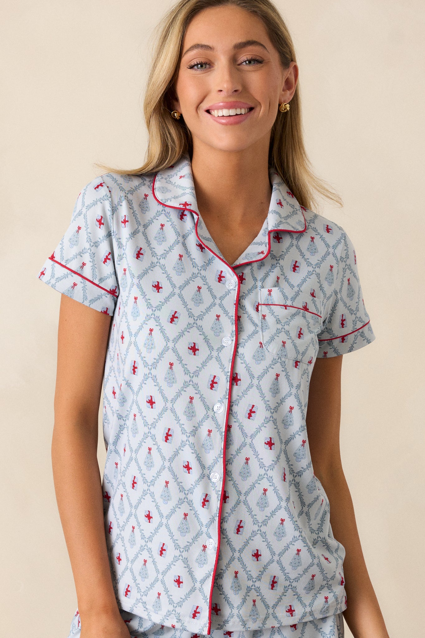 Untucked front view of a light blue pajama shirt featuring a collared neckline, red trim, a front pocket, a beautiful tree and present holiday print, a relaxed fit and short sleeves.