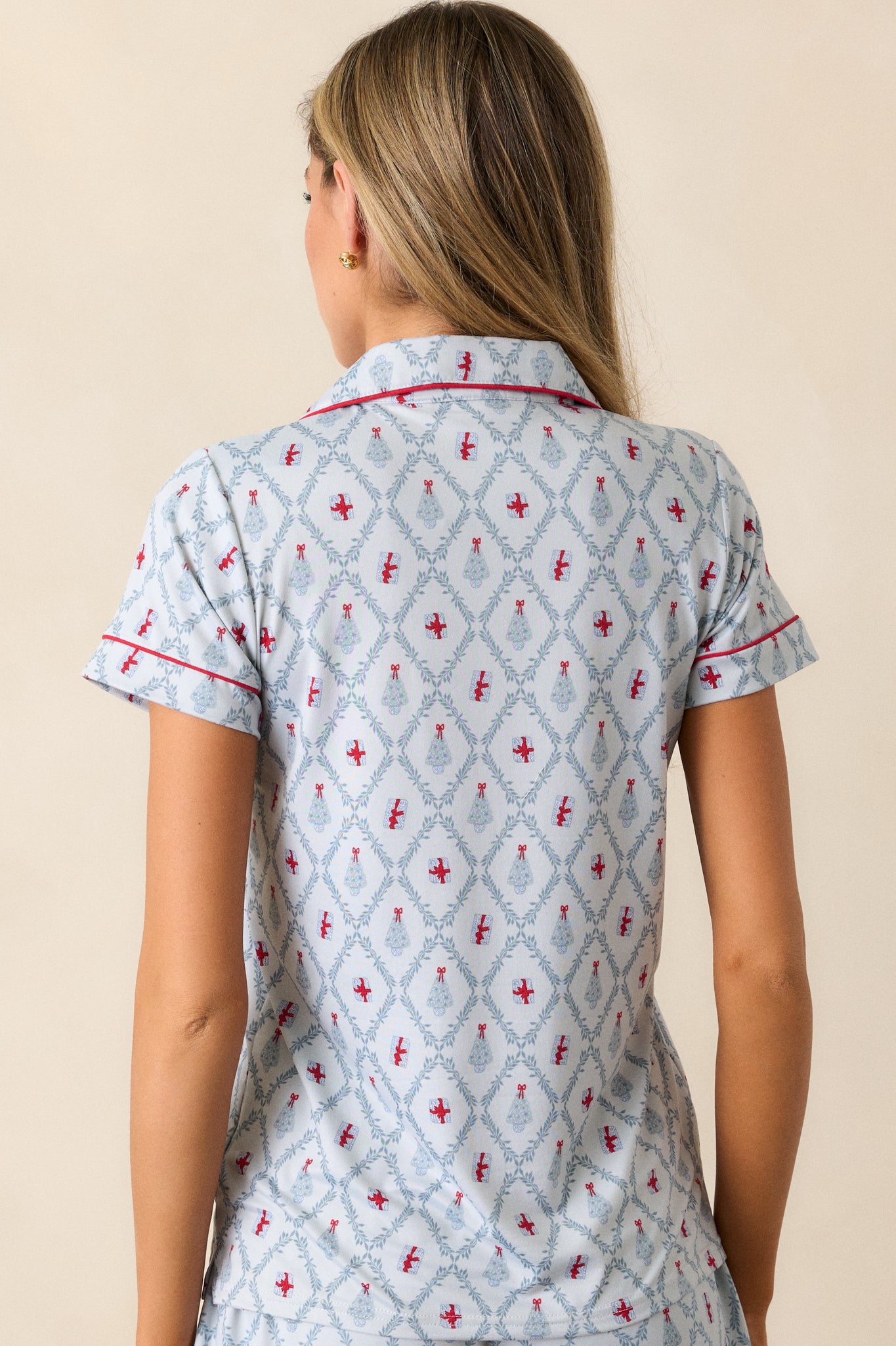 Back view of a light blue pajama shirt featuring a collared neckline, red trim, a front pocket, a beautiful tree and present holiday print, a relaxed fit and short sleeves.