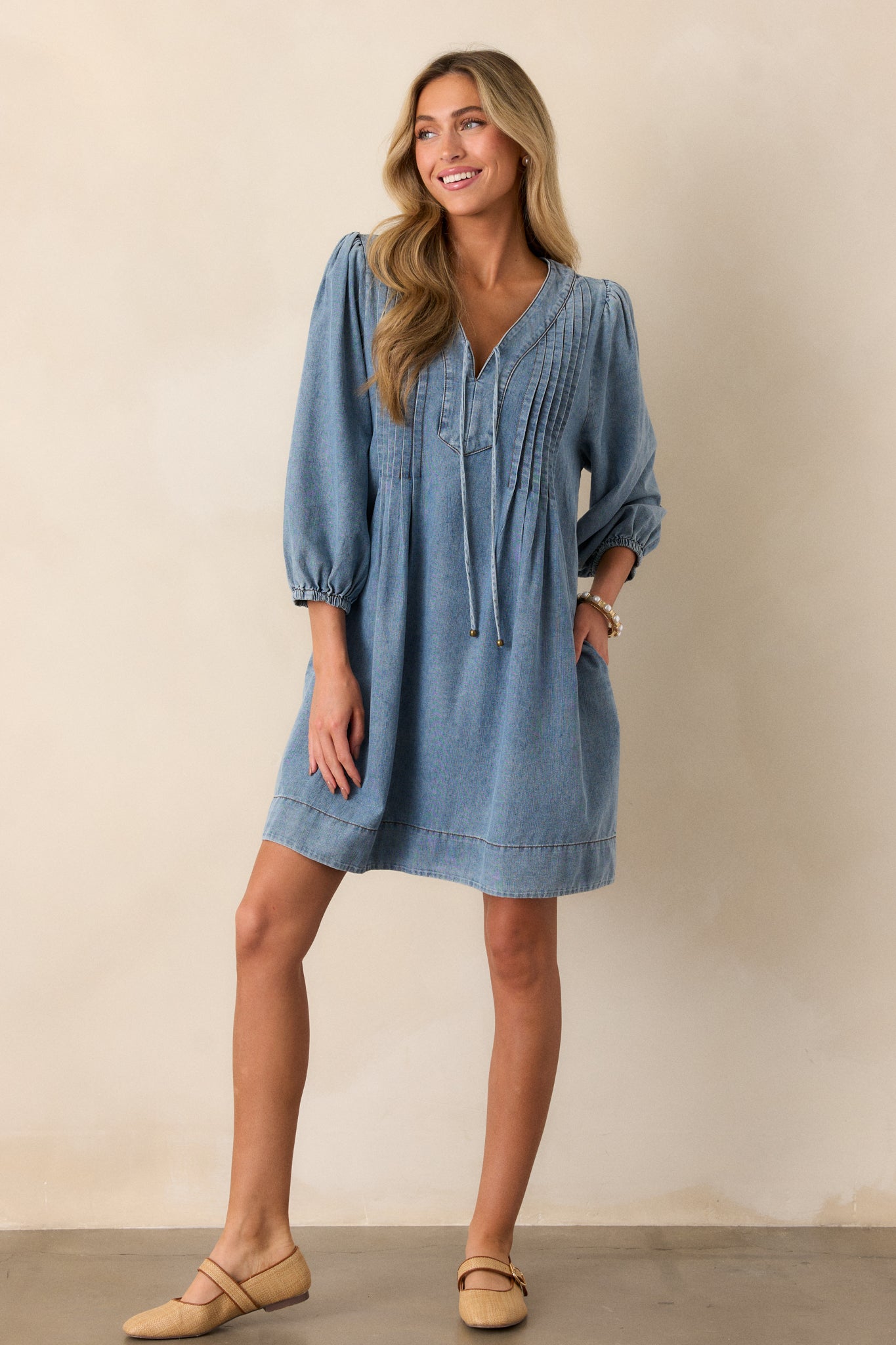 A front-facing view of the light wash denim dress, highlighting the pleated detailing and v-neckline alongside the long sleeves.