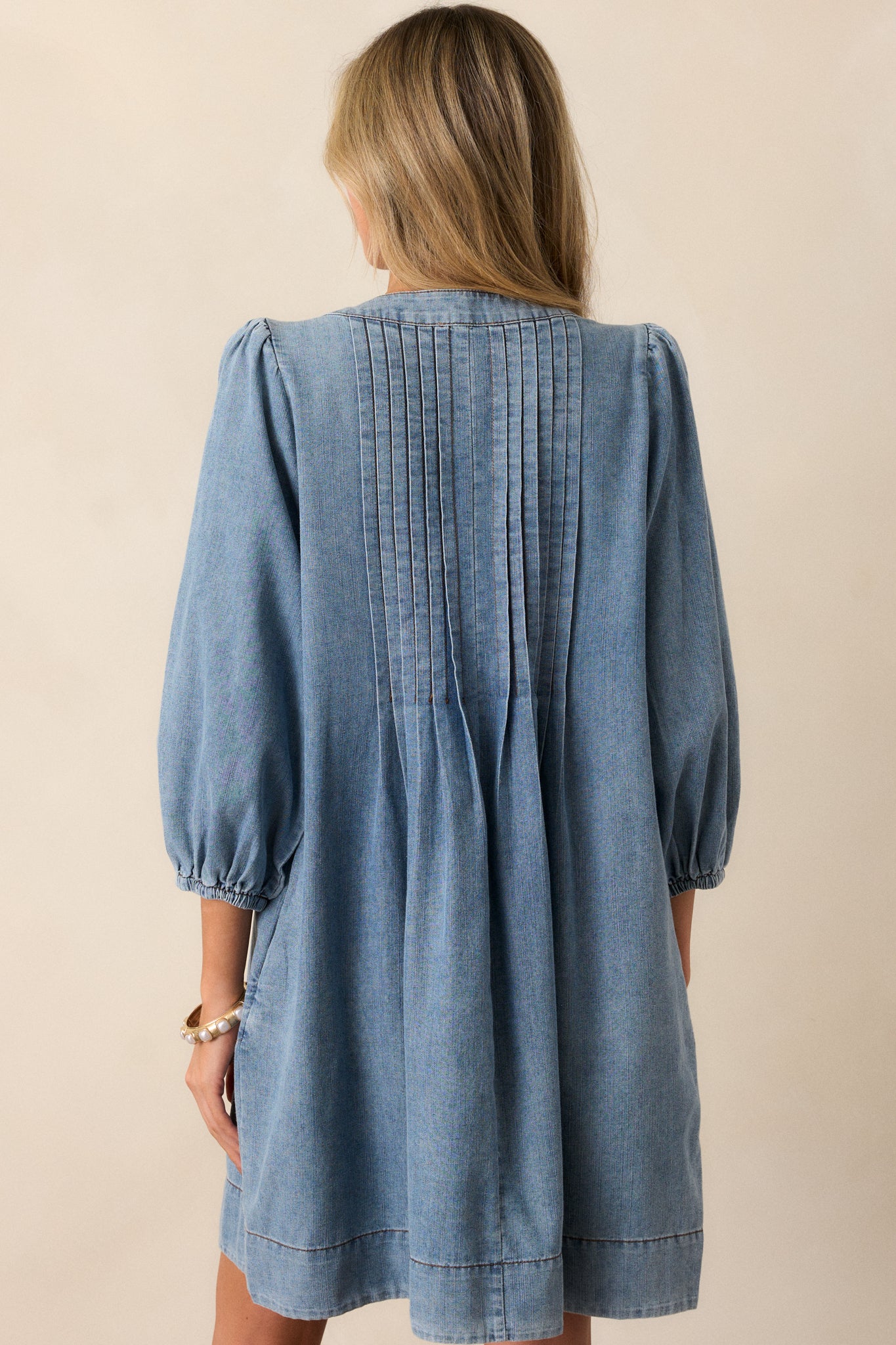 A back view of the light wash denim dress, showcasing the long sleeves and the pleated detailing along the back.