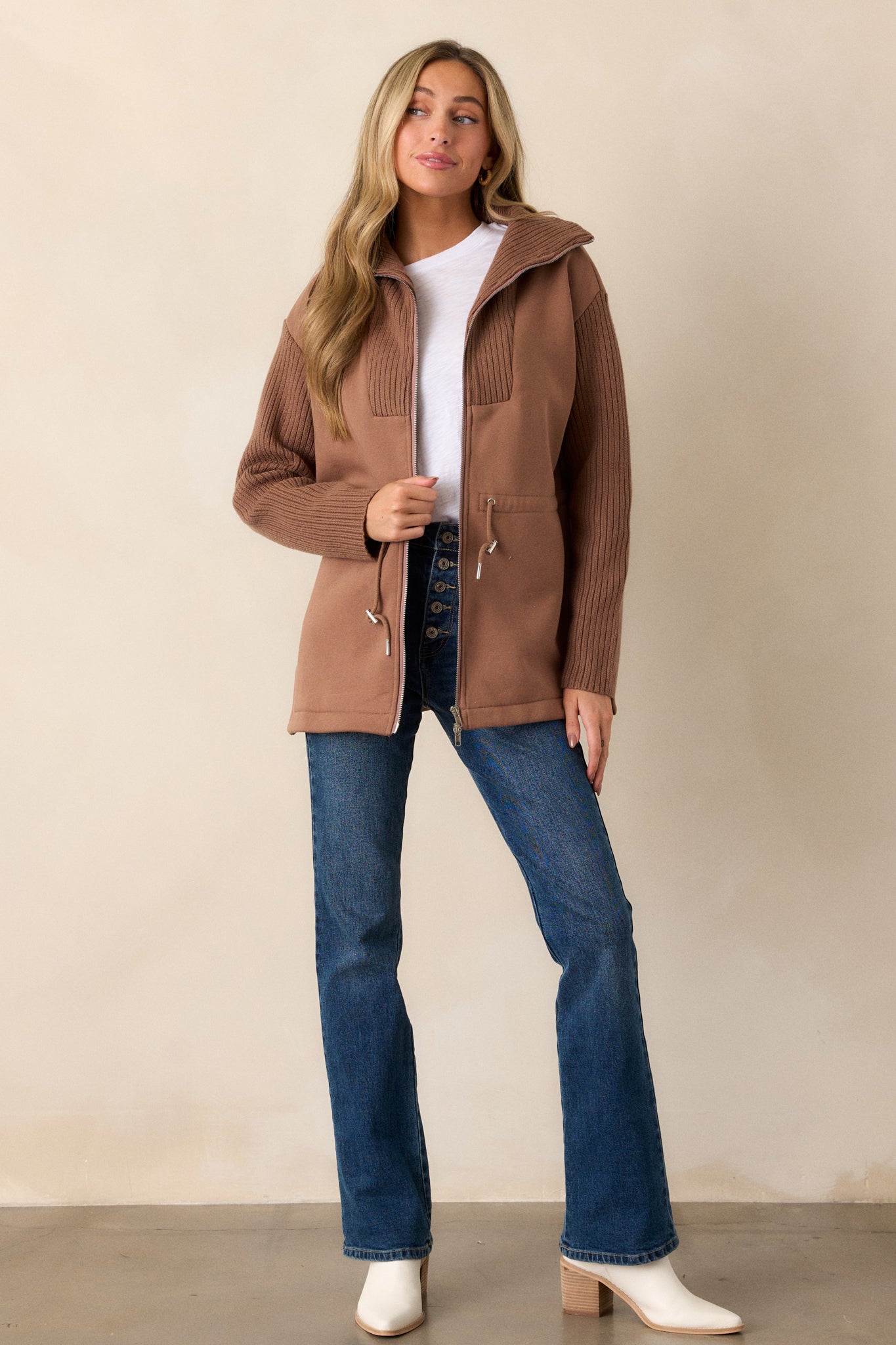 A full-body shot of the brown jacket, showcasing the knit high neck, front zipper, and the fitted drawstring waistband with silver details.