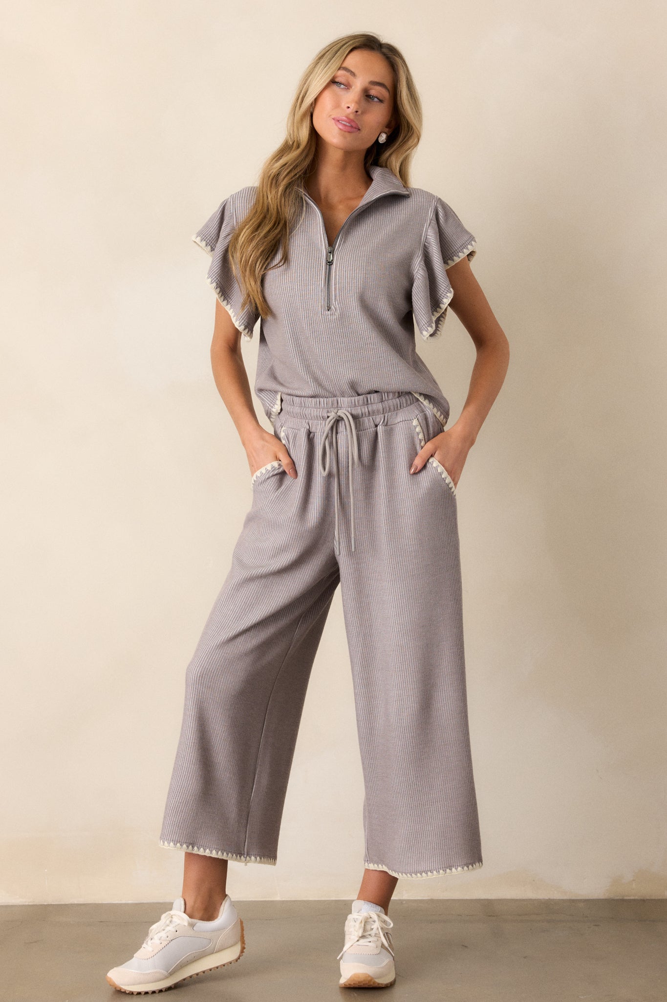 These grey pants feature an elastic waistband, an adjustable drawstring, ivory blanket stitch detailing, functional hip pockets, a soft knit material, and a wide leg.