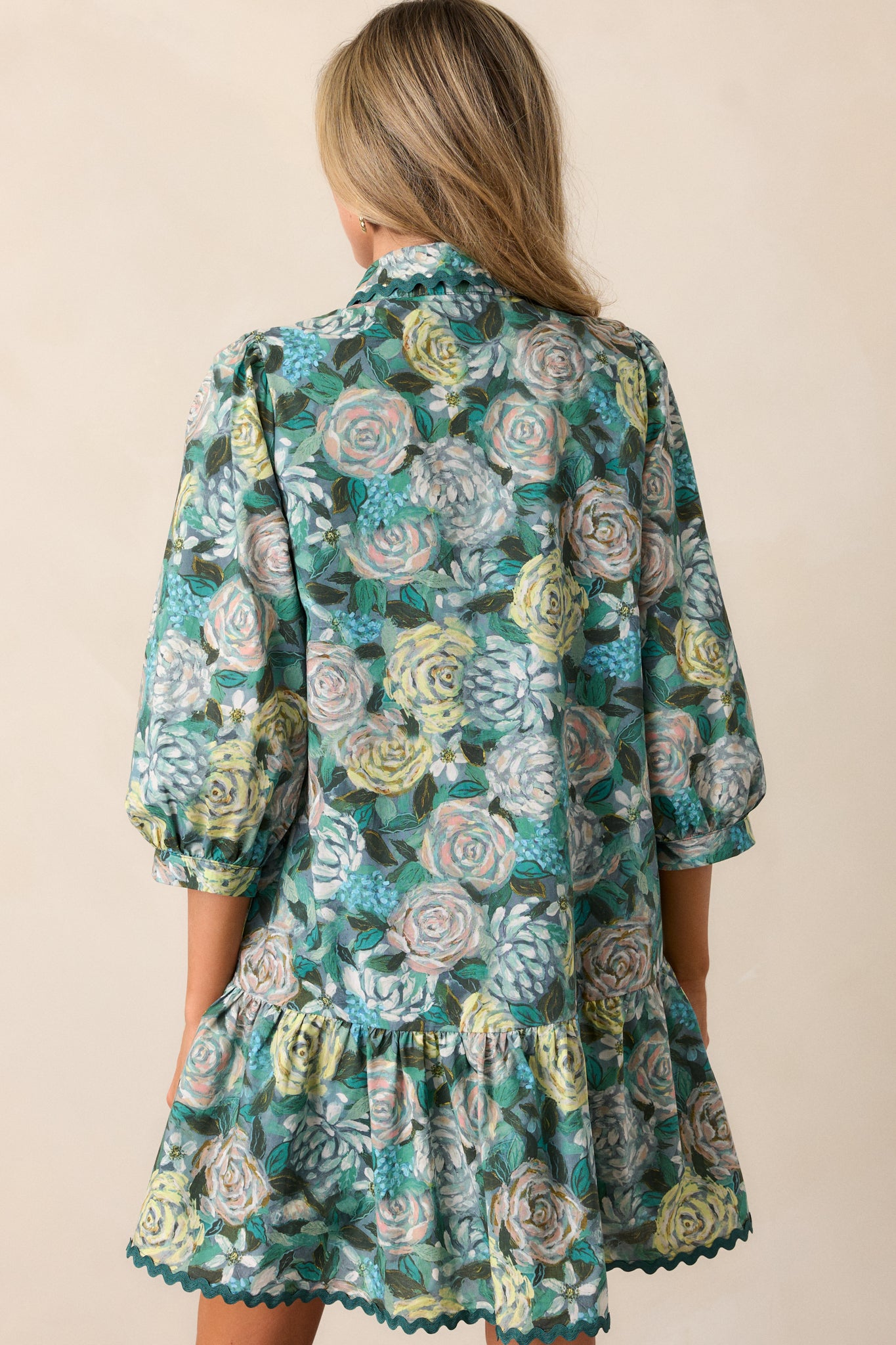 A back view of the green mini dress, showcasing the full floral pattern, tiered bottom, and 3/4 sleeves. The overall structure and relaxed fit of the dress are also visible.