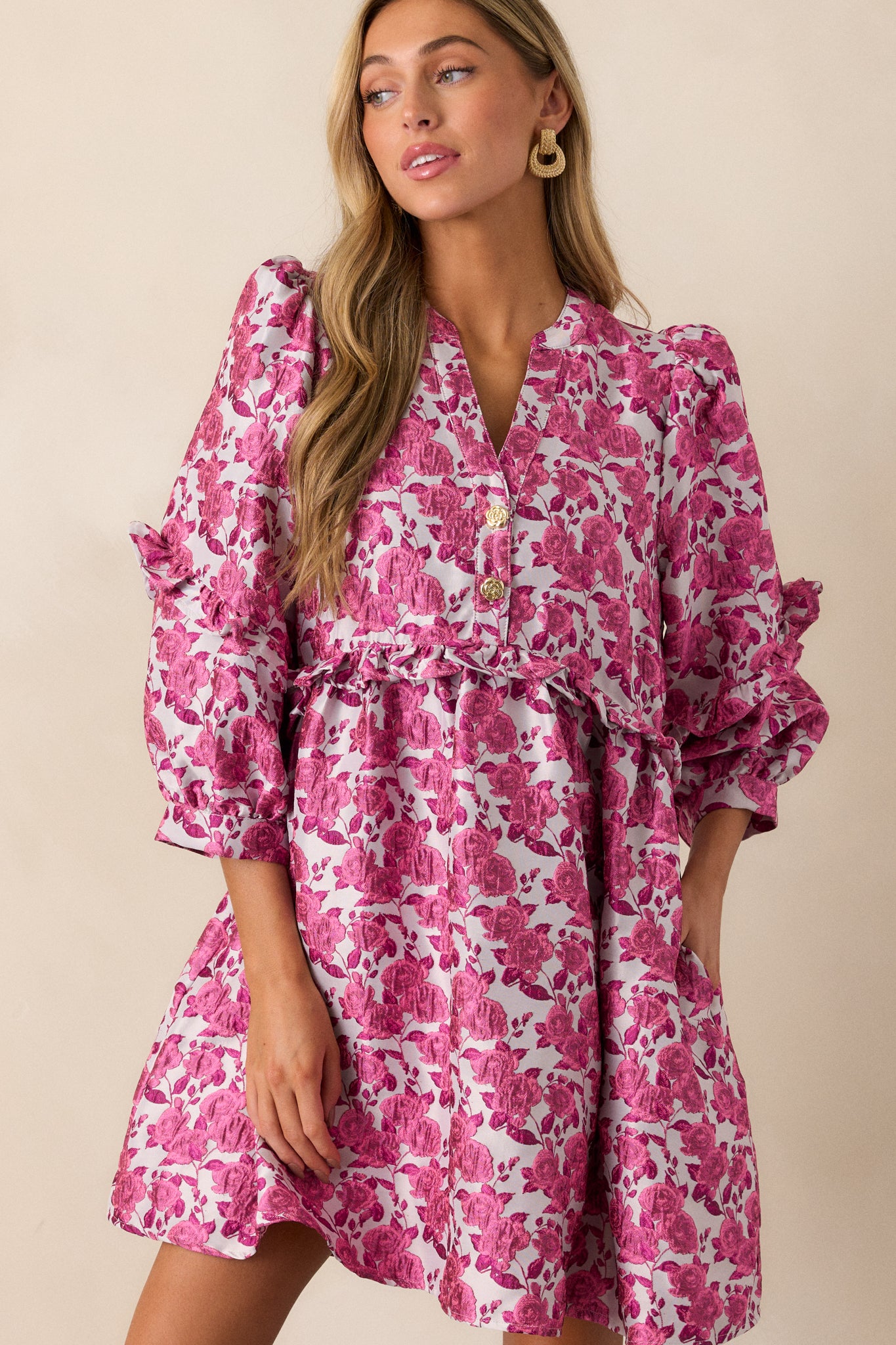 Front view of a pink dress featuring floral pattern, a slight v-neckline with functional gold floral embellished buttons. a ruffle waistline, and 3/4 length puff sleeves with ruffle detailing.