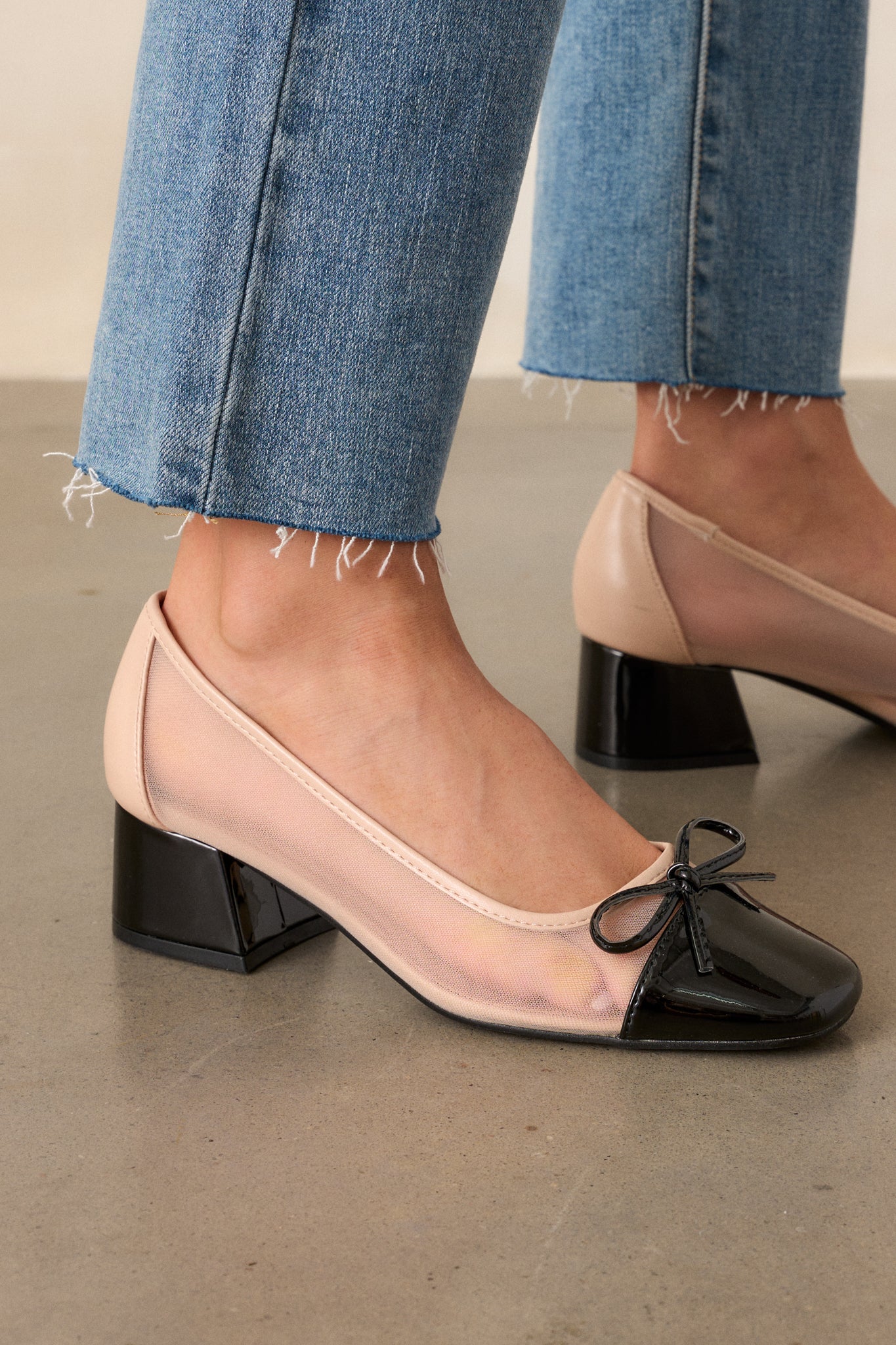 A cropped view of the beige and black heels, highlighting the glossy black square toe and the slim black bow accent on the toe.