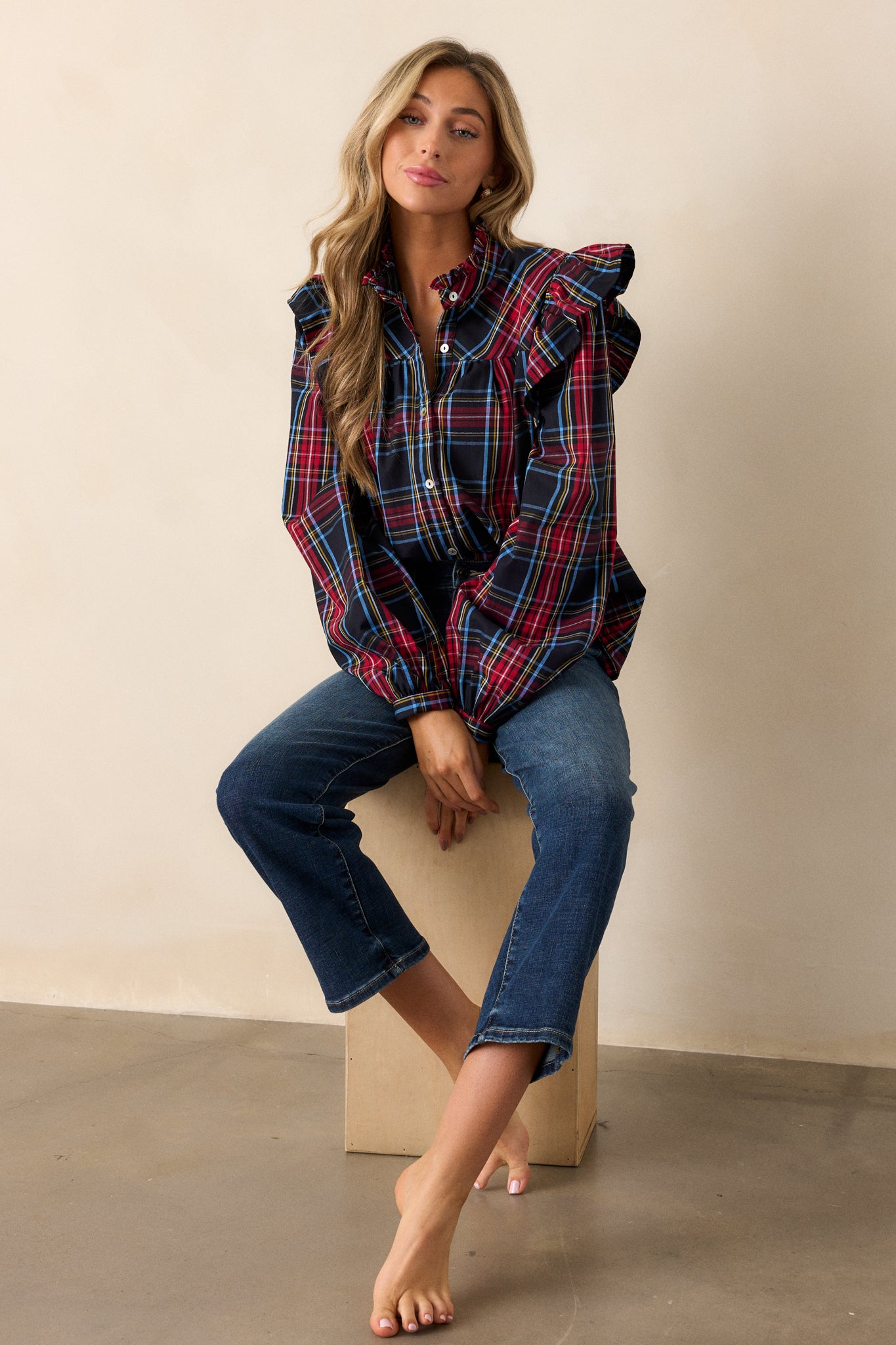 Seated full body view of this black plaid top featuring a ruffle round neckline, ruffle sleeve detail, functional buttons down the center, wide sleeves, and buttoned cuffs.  