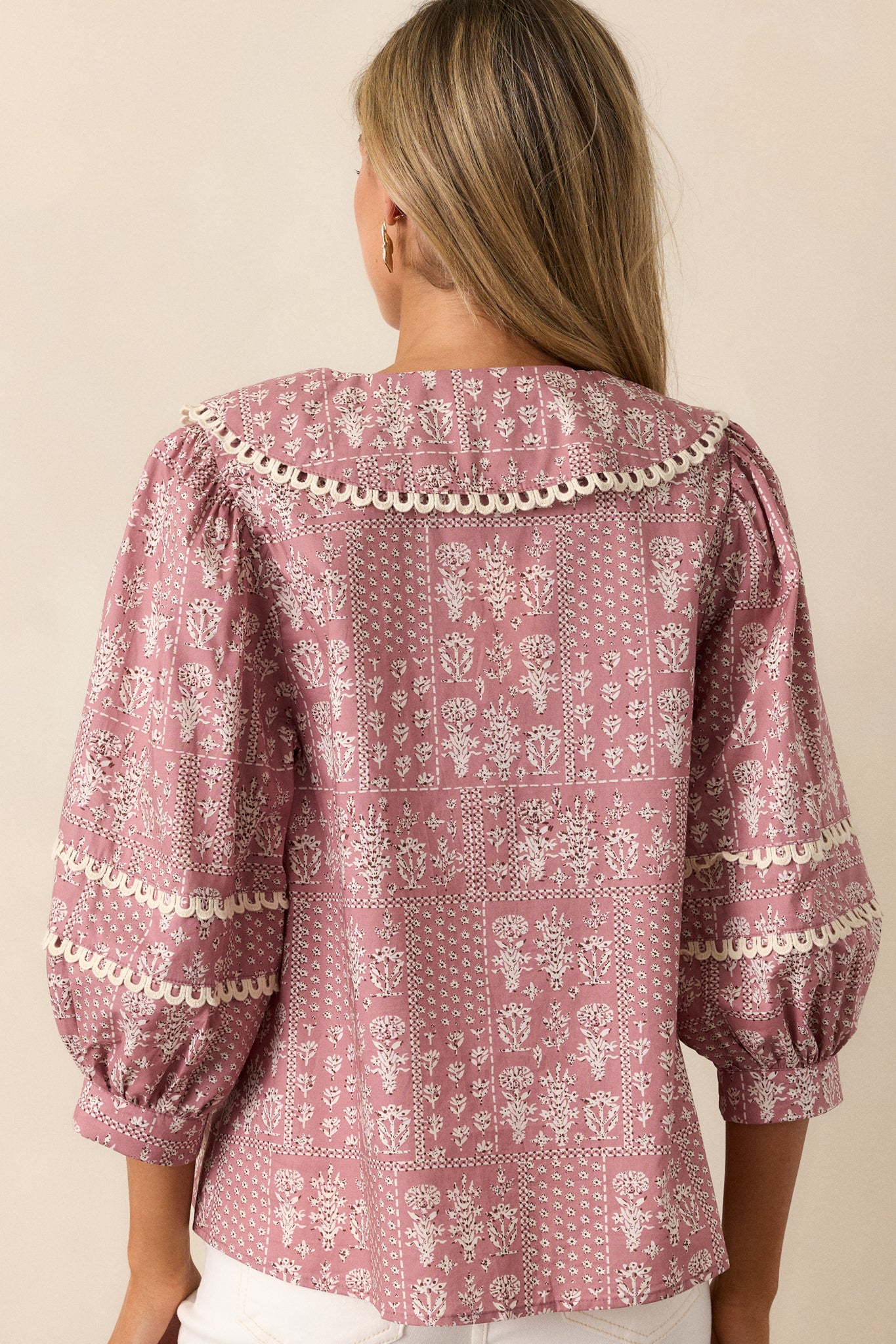 Back view of a dusty pink blouse featuring a collared v-neckline, button up front, small scalloped hemming, a beautiful floral pattern, and 3/4 puff sleeves with scalloped detailing.