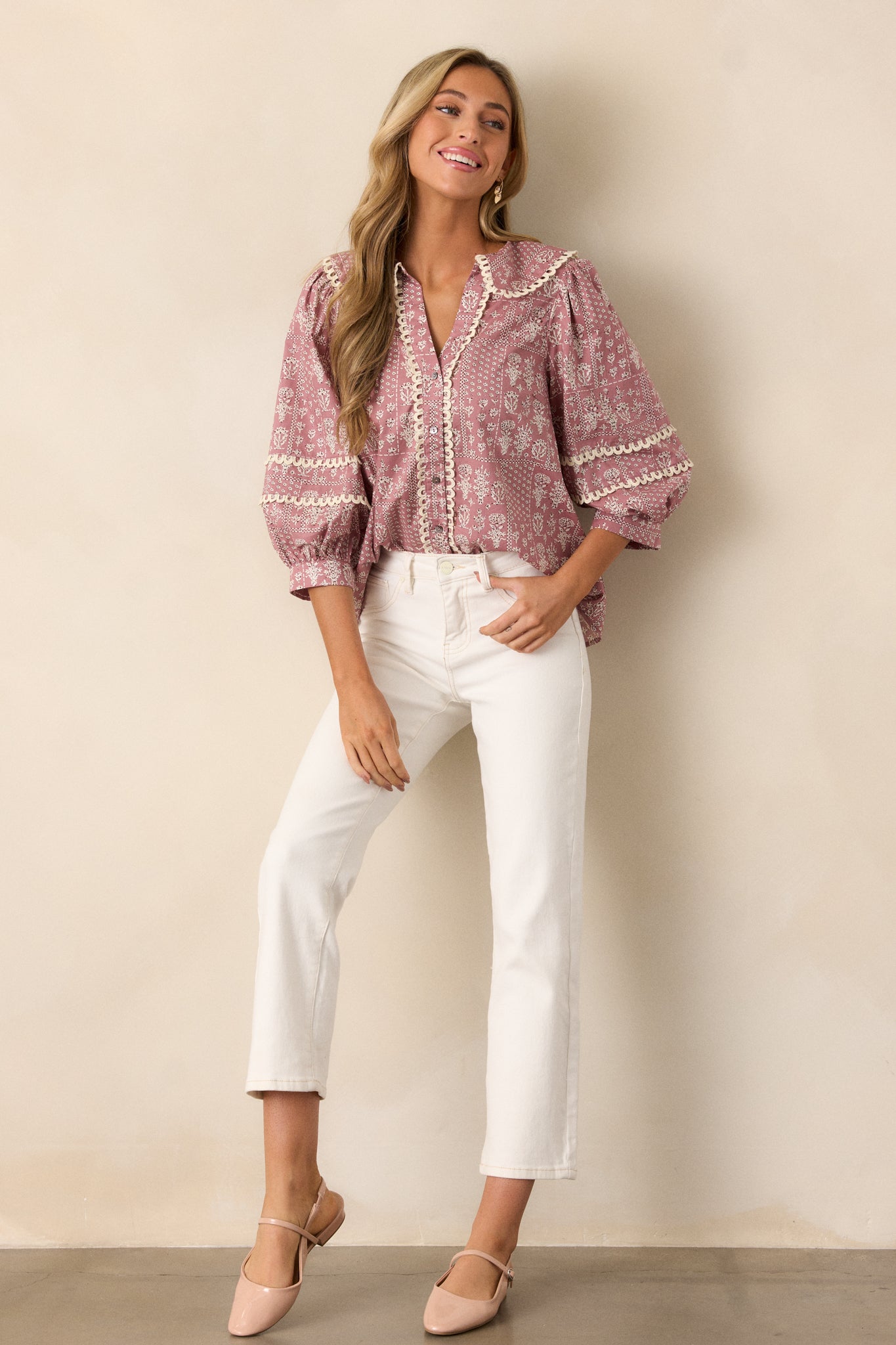 Posed view of a dusty pink blouse featuring a collared v-neckline, button up front, small scalloped hemming, a beautiful floral pattern, and 3/4 puff sleeves with scalloped detailing.