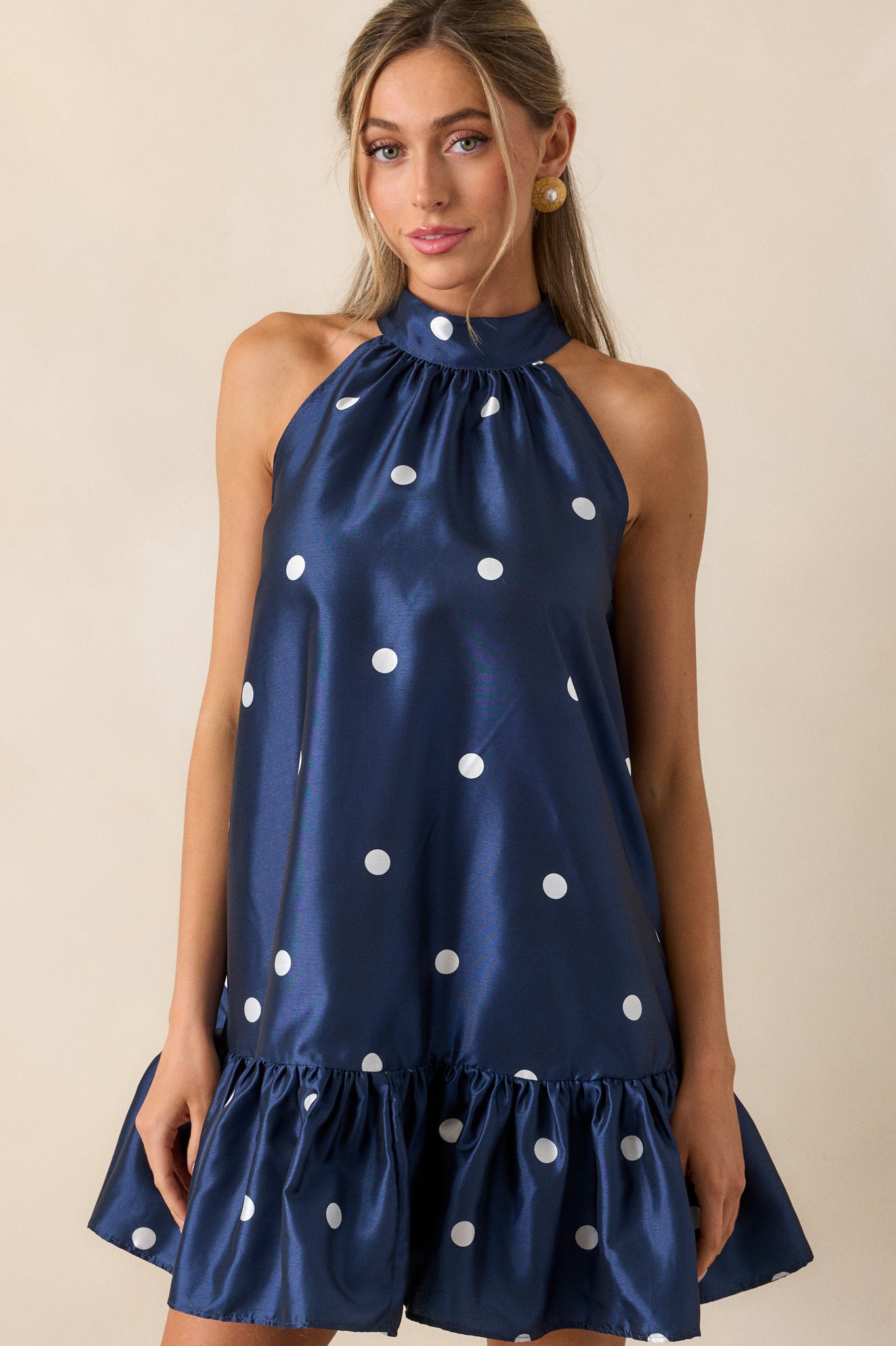 A full-length shot of the navy mini dress featuring a halter neckline with button clasps. The dress has a babydoll cut with a ruffled bottom hem, showcasing the polka dot pattern throughout.