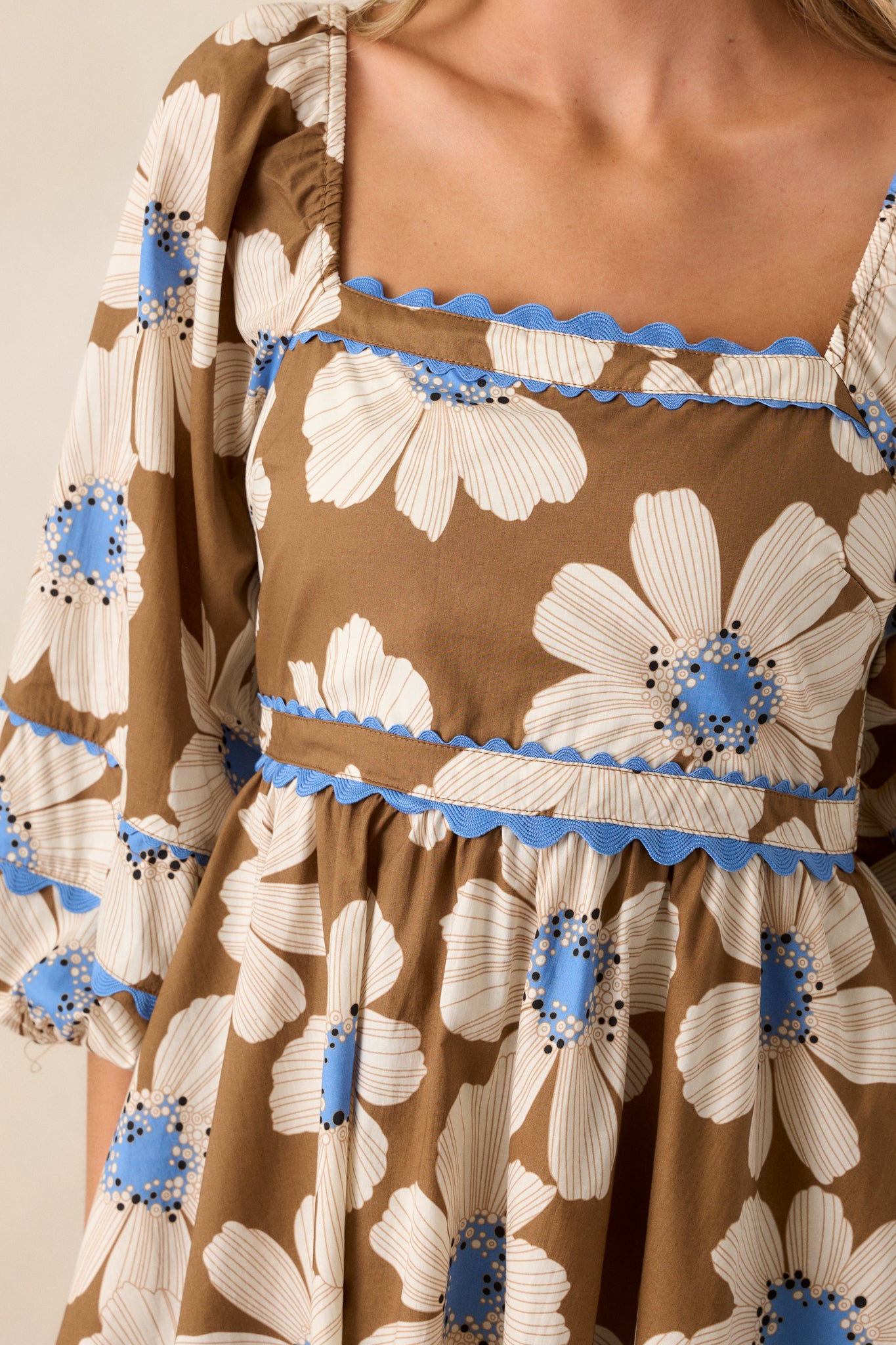 Close-up shot of the blue ric rac trim along the neckline, puff sleeve cuffs, and bottom hem, highlighting the floral print in ivory and light blue. The smocked back insert adds texture and shape.