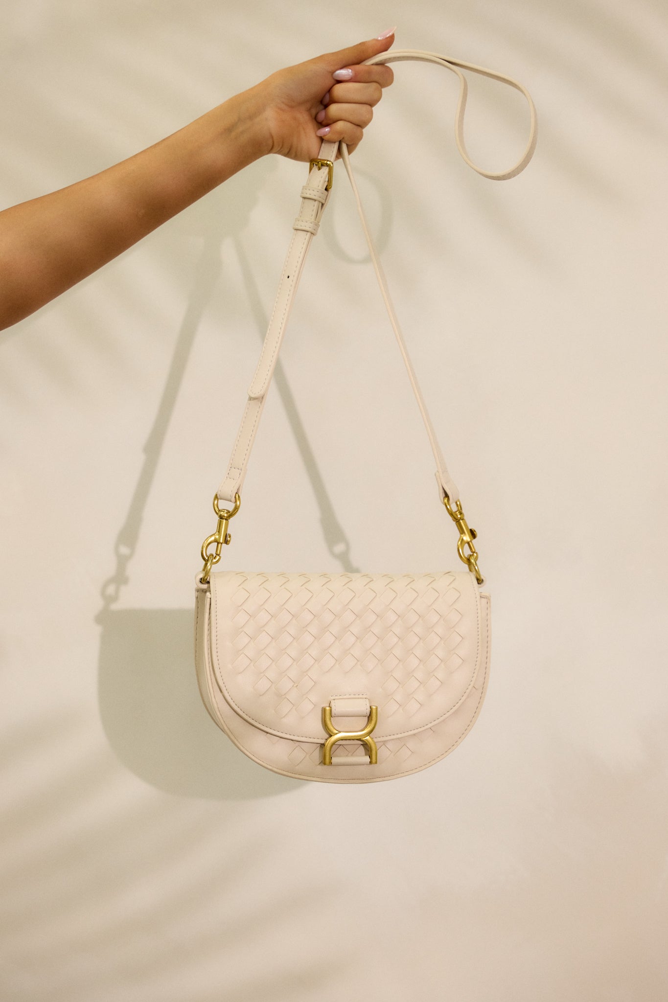 A cropped image showing the gold hardware and fold-over design of the beige handbag. The solid beige shoulder strap, resembling a belt, adds to the unique style of the handbag.