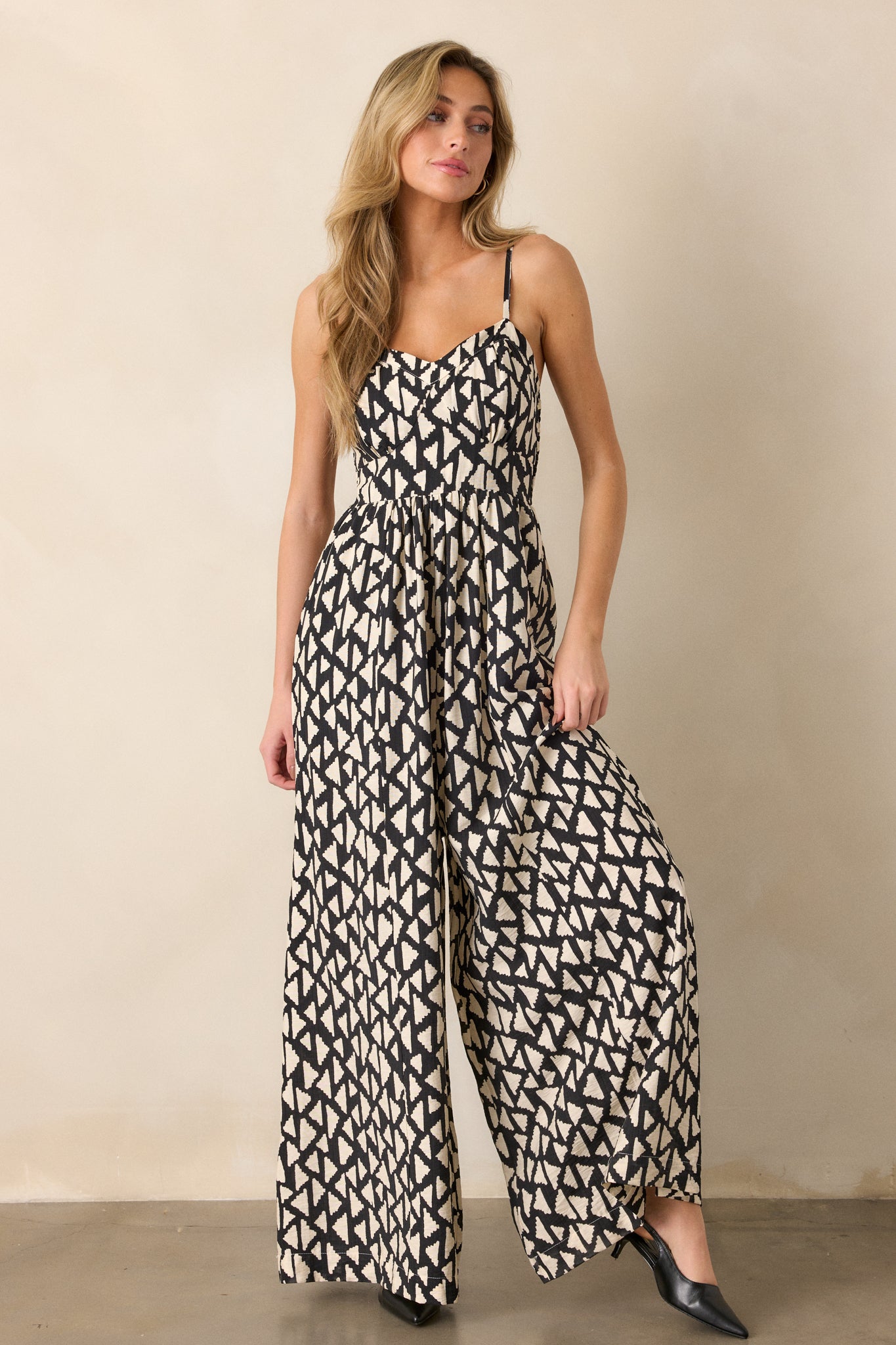 A black jumpsuit with a geometric print and adjustable straps is displayed in a full-body view from another angle.