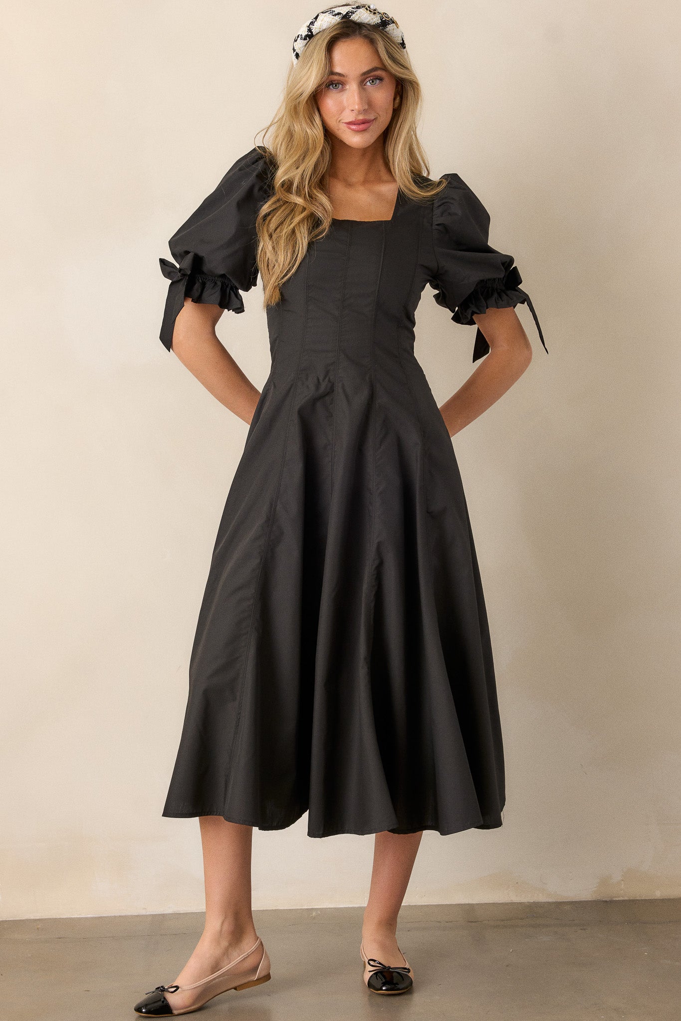 A black midi dress with a square neckline and a fit-and-flare silhouette, featuring short puff sleeves with bow accents and smocked details.