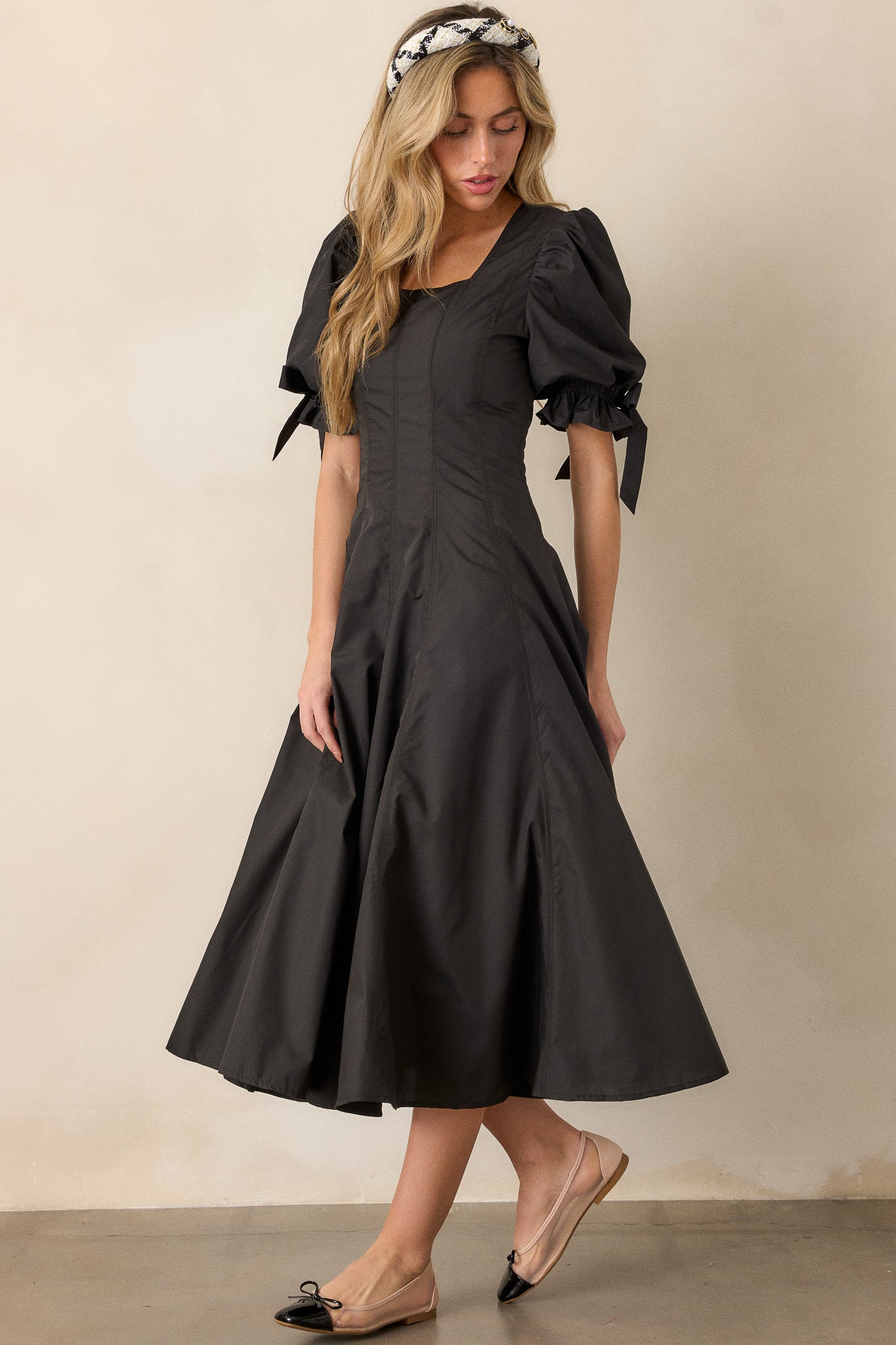 An angled shot of the black dress, emphasizing the fit-and-flare structure, square neckline, and bow accents on the short puff sleeves.