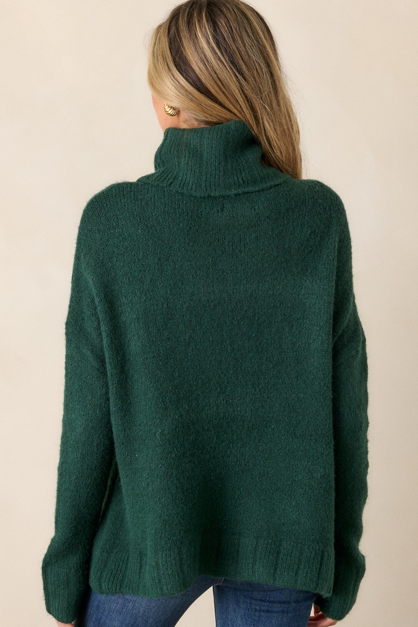 Back view of this sweater that features a very light eyelash knit design, a turtle cowl neckline, extremely dropped shoulders, and a slight high low hemline.