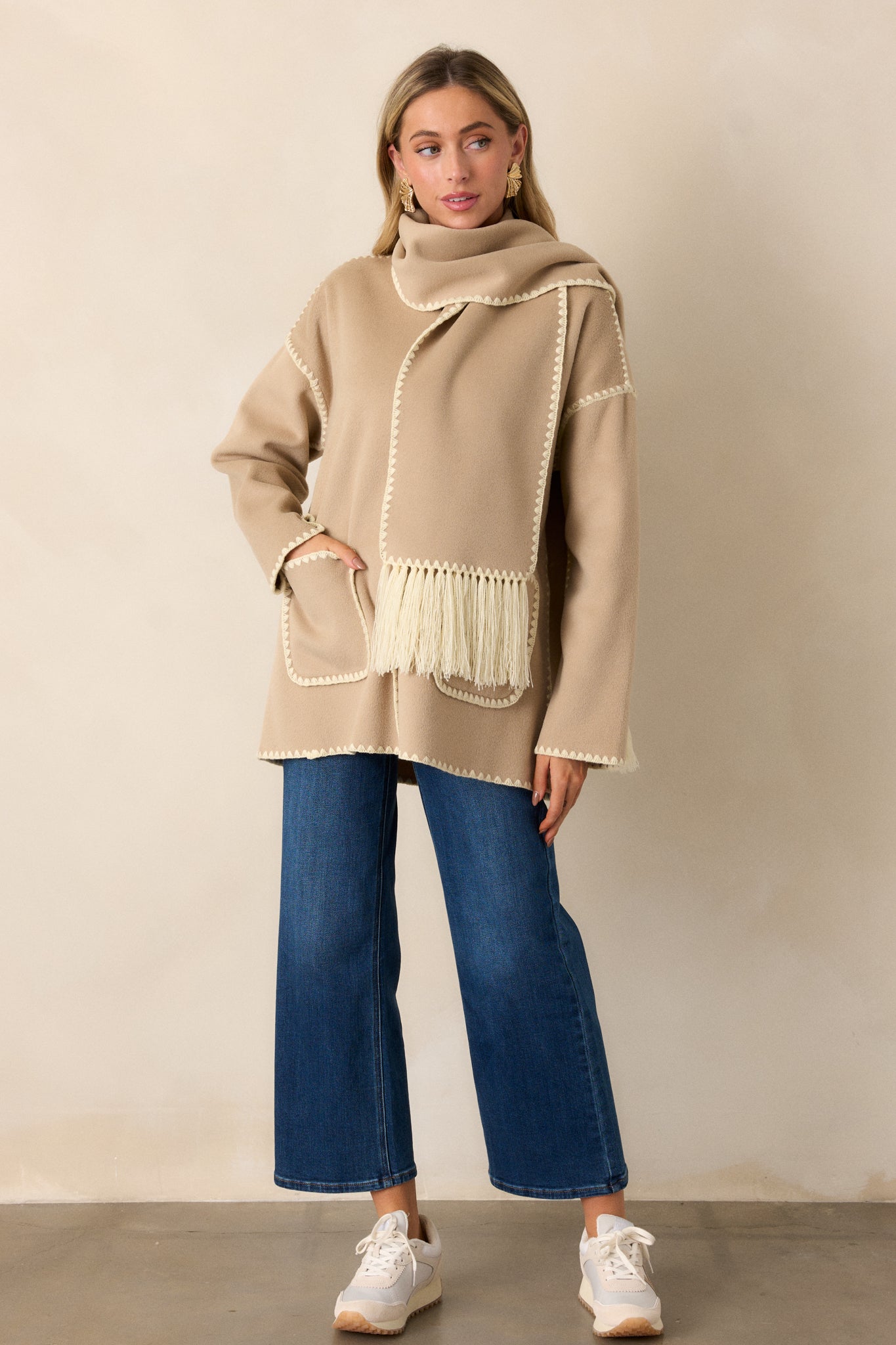 Full body view of a Taupe Coat featuring a rounded neckline, functional button front, blanket stitch hems, functional front pockets, long sleeves with blanket stitch detailing and a blanket stitch scarf with fringe detailing.