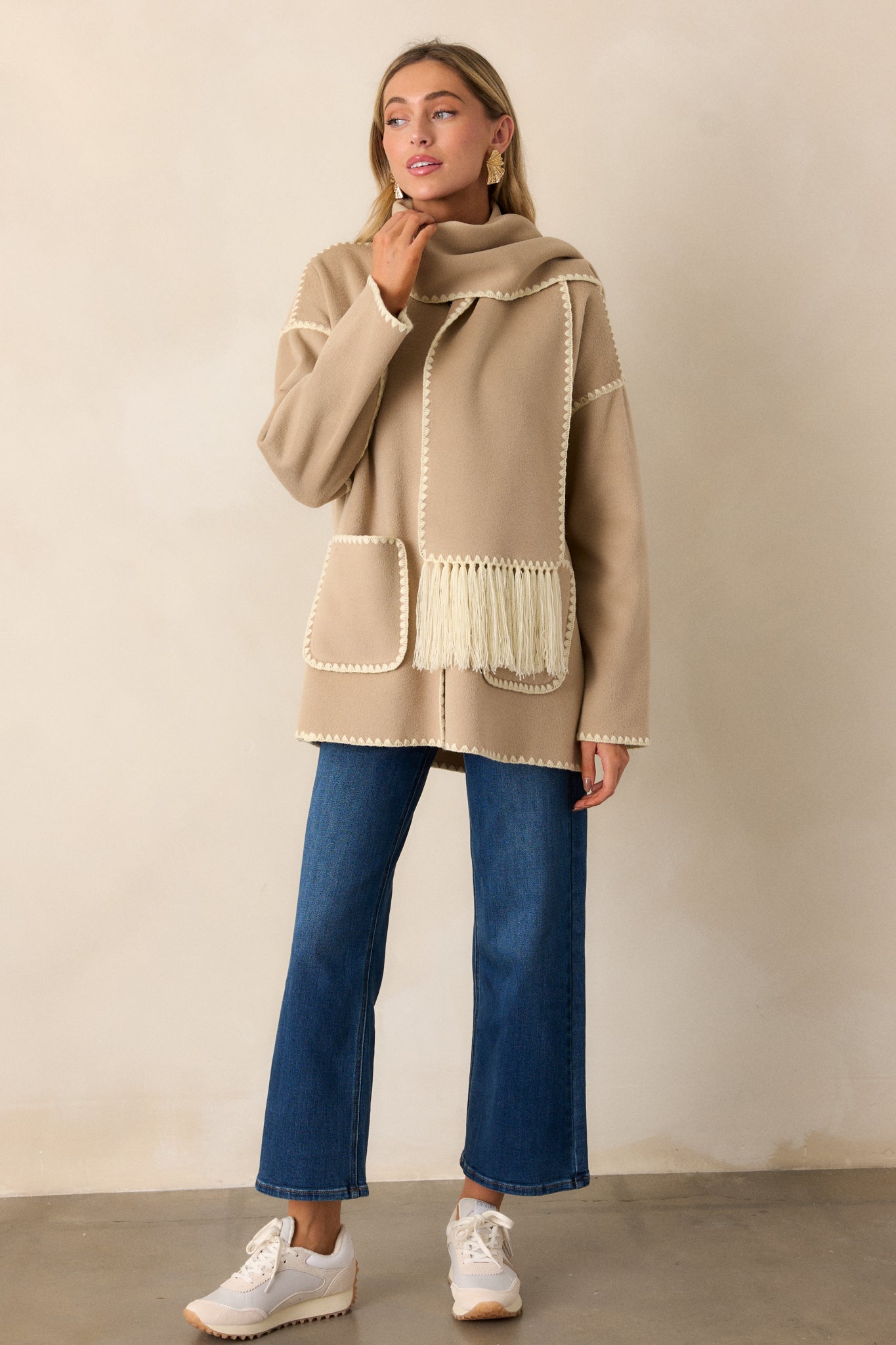 Fully buttoned up view of a taupe coat with blanket stitch detailing, dropped shoulders, functional pockets, functional buttons, and a matching scarf with fringe detailing. 