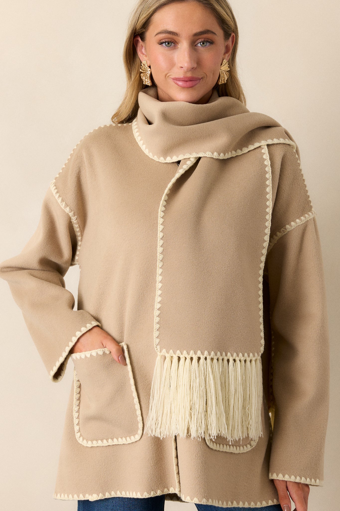 Front view of a Taupe Coat featuring a rounded neckline, functional button front, blanket stitch hems, functional front pockets, long sleeves with blanket stitch detailing and a blanket stitch scarf with fringe detailing.