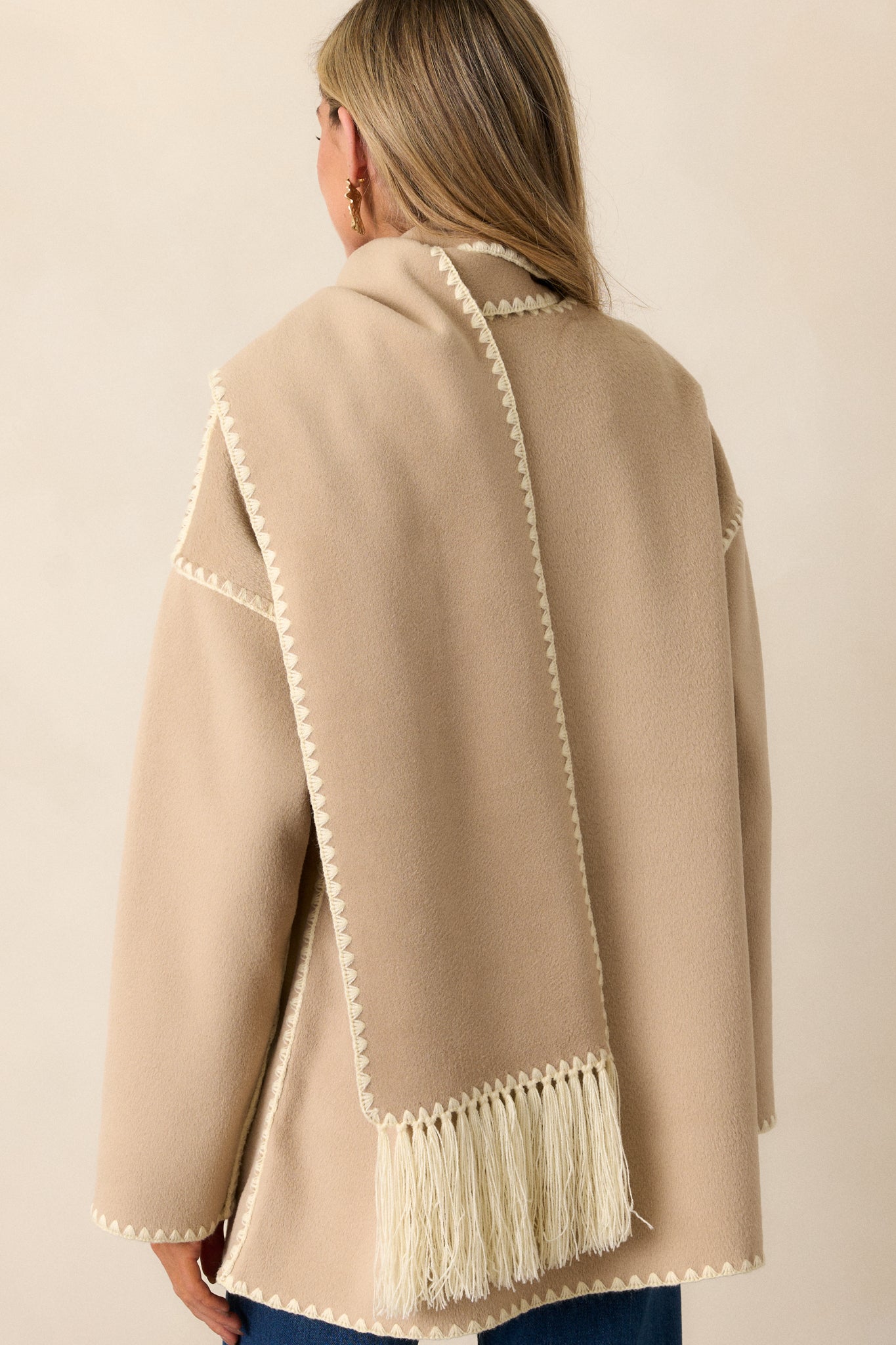 Back view of a Taupe Coat featuring a rounded neckline, functional button front, blanket stitch hems, functional front pockets, long sleeves with blanket stitch detailing and a blanket stitch scarf with fringe detailing.