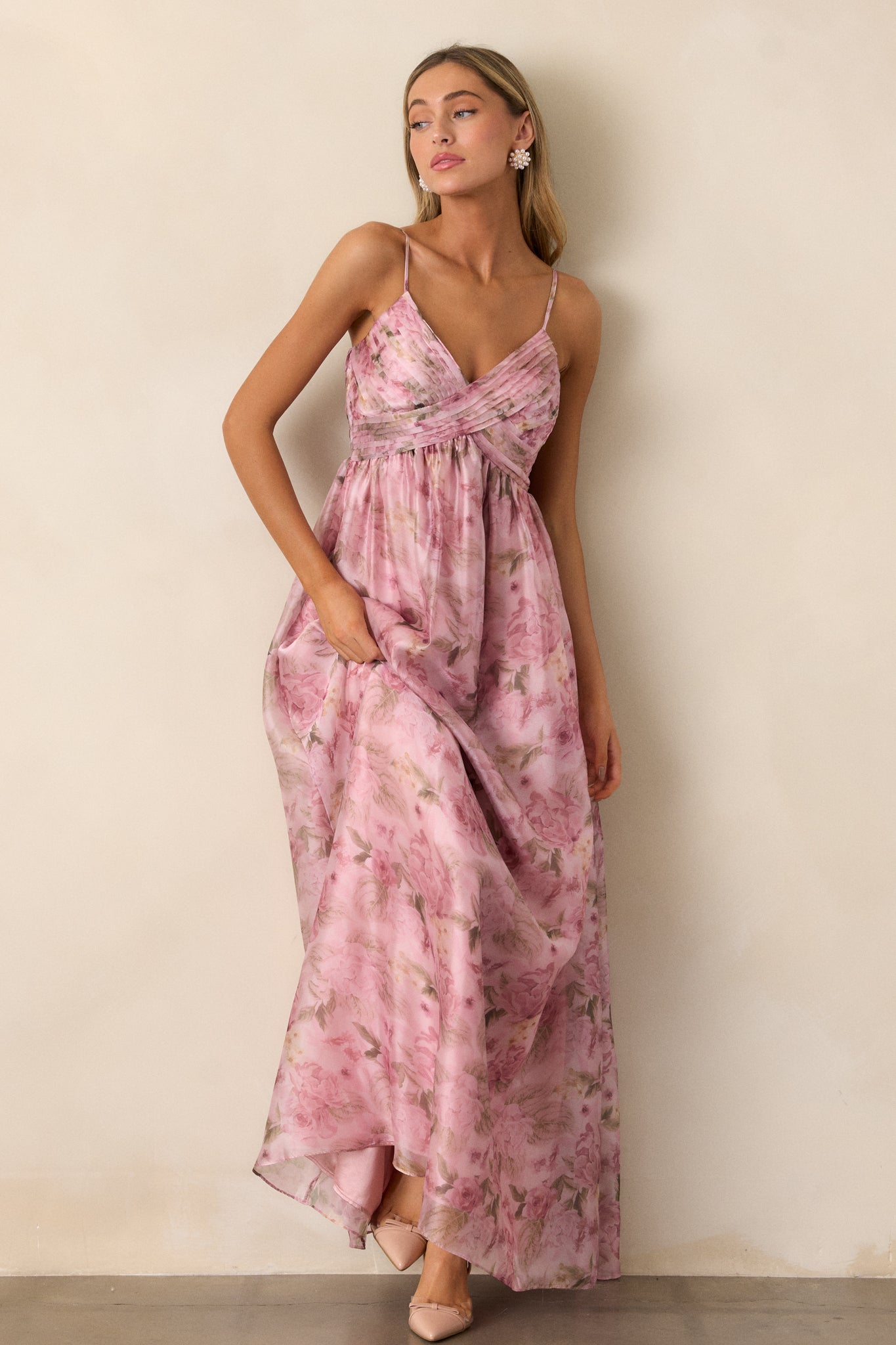 Full body view of a pink dress featuring a v- neckline, empire waist, cross pleated detailing on the top, flowing skirt, an elegant floral design throughout, and spaghetti straps.