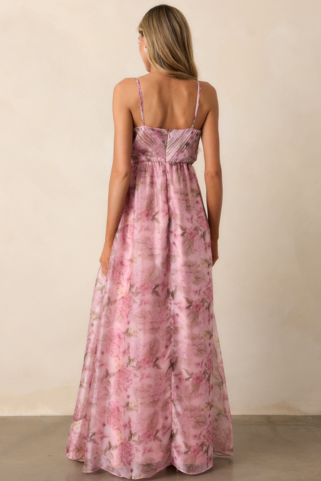 Back view of a pink dress featuring a v- neckline, empire waist, cross pleated detailing on the top, flowing skirt, an elegant floral design throughout, and spaghetti straps.