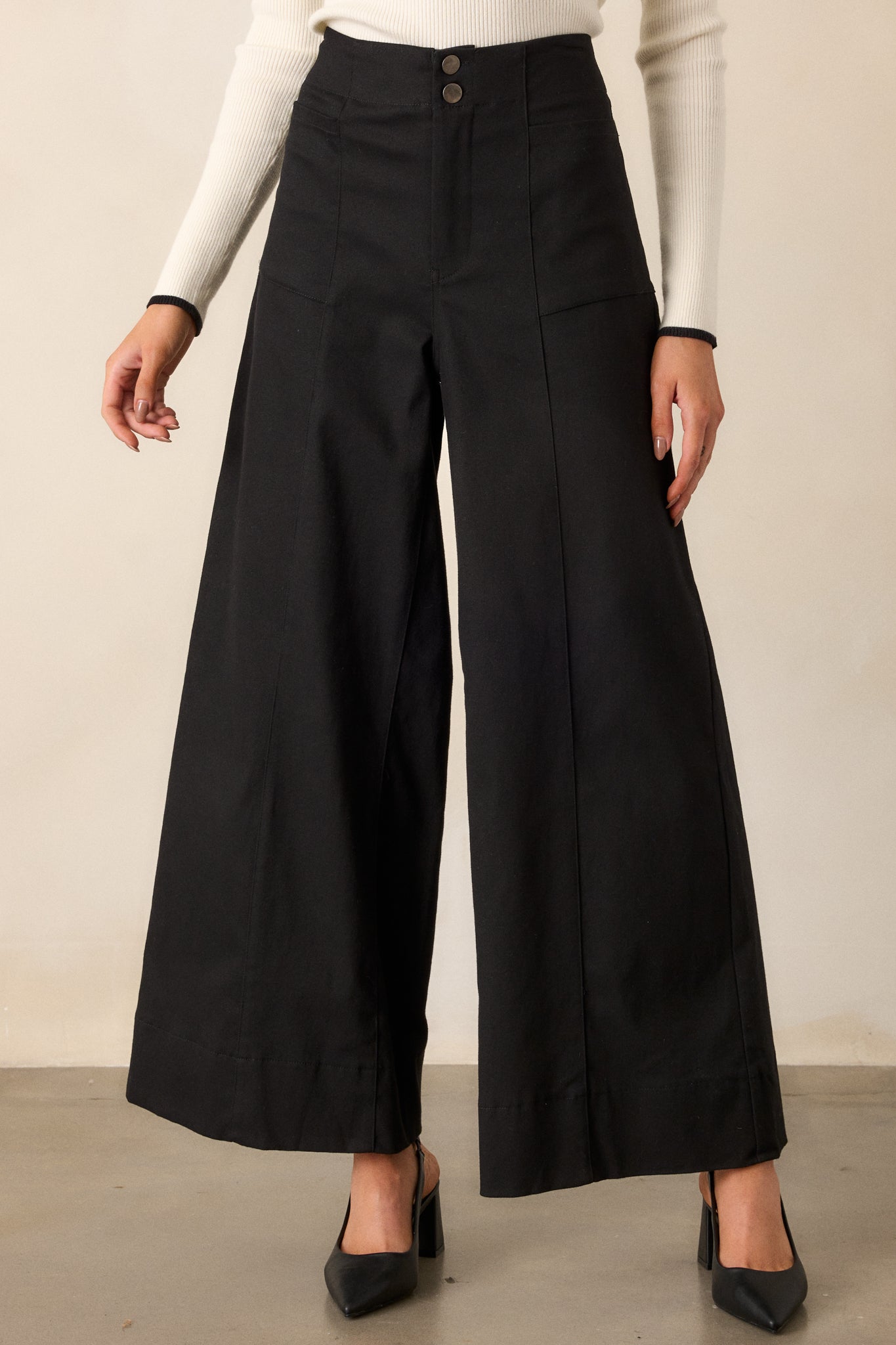 A zoomed-in view of black pants featuring a high waisted design, a button zipper closure, functional rectangular hip pockets, and a wide leg.