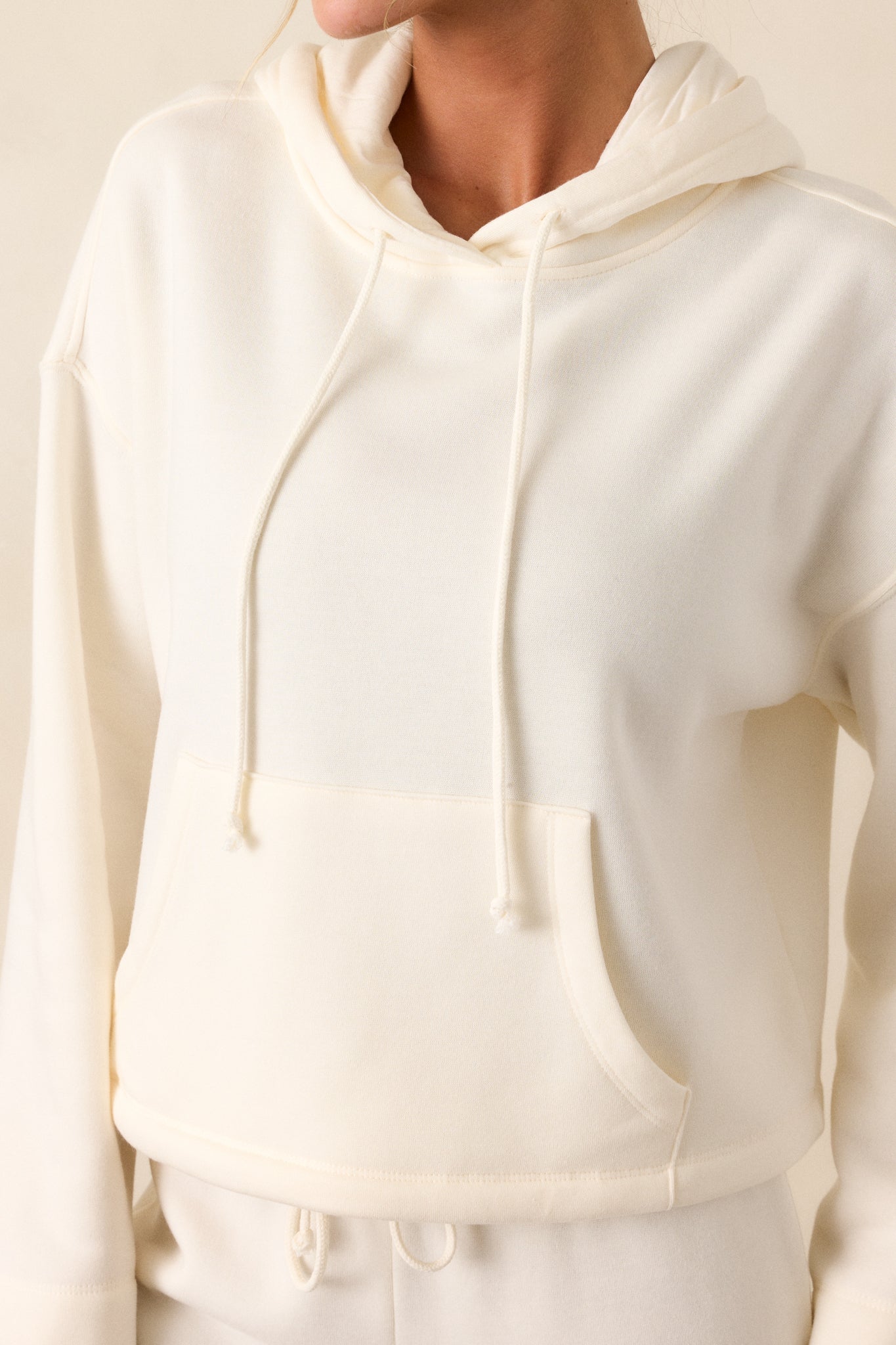 Close-up of the ivory sweatshirt’s fabric texture, focusing on the adjustable drawstring and stitching around the kangaroo pocket.