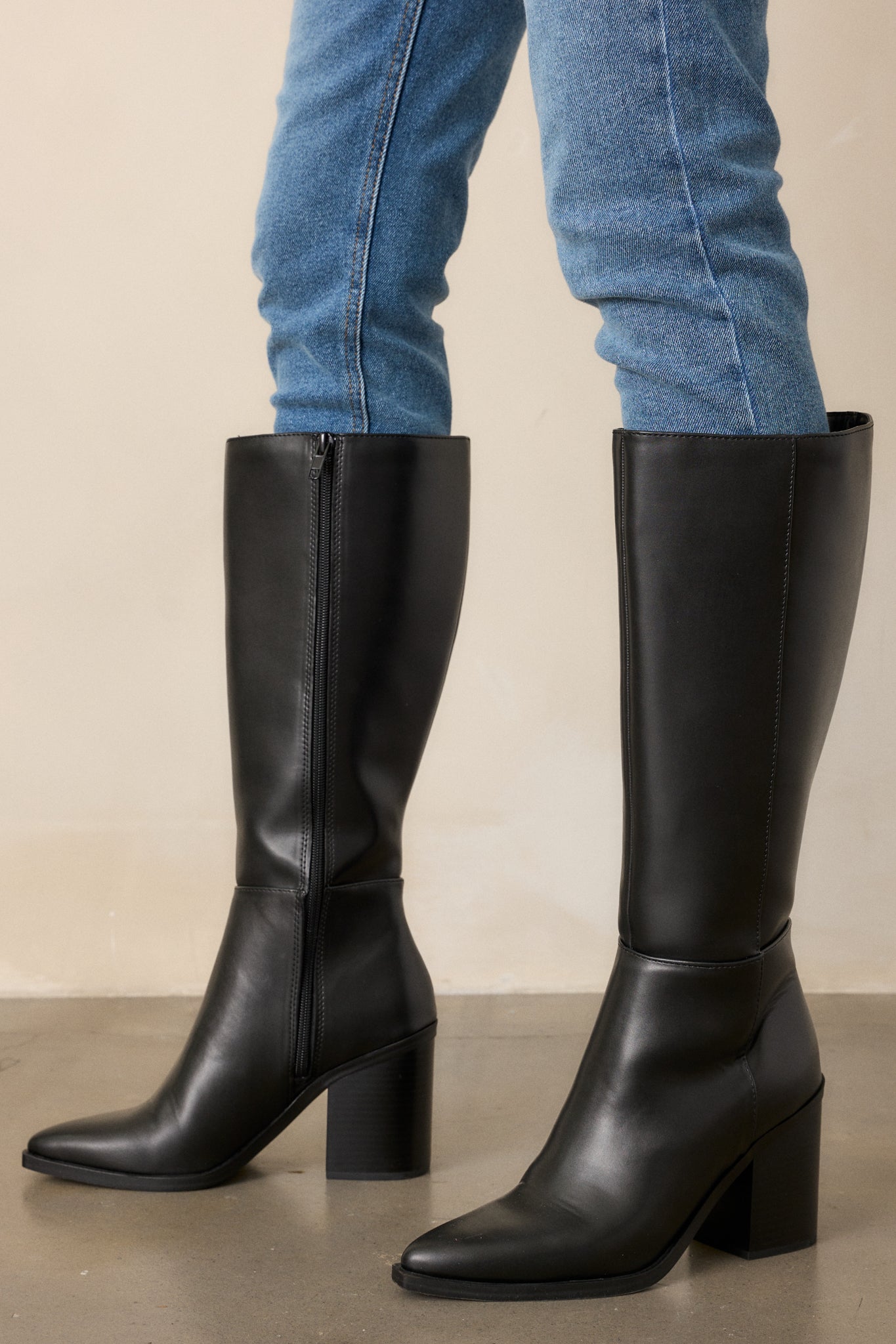 Side view of black boots featuring a pointed toe, a sleek faux leather black material, a black block heel, a functional side zipper, a knee high design and a black sole.