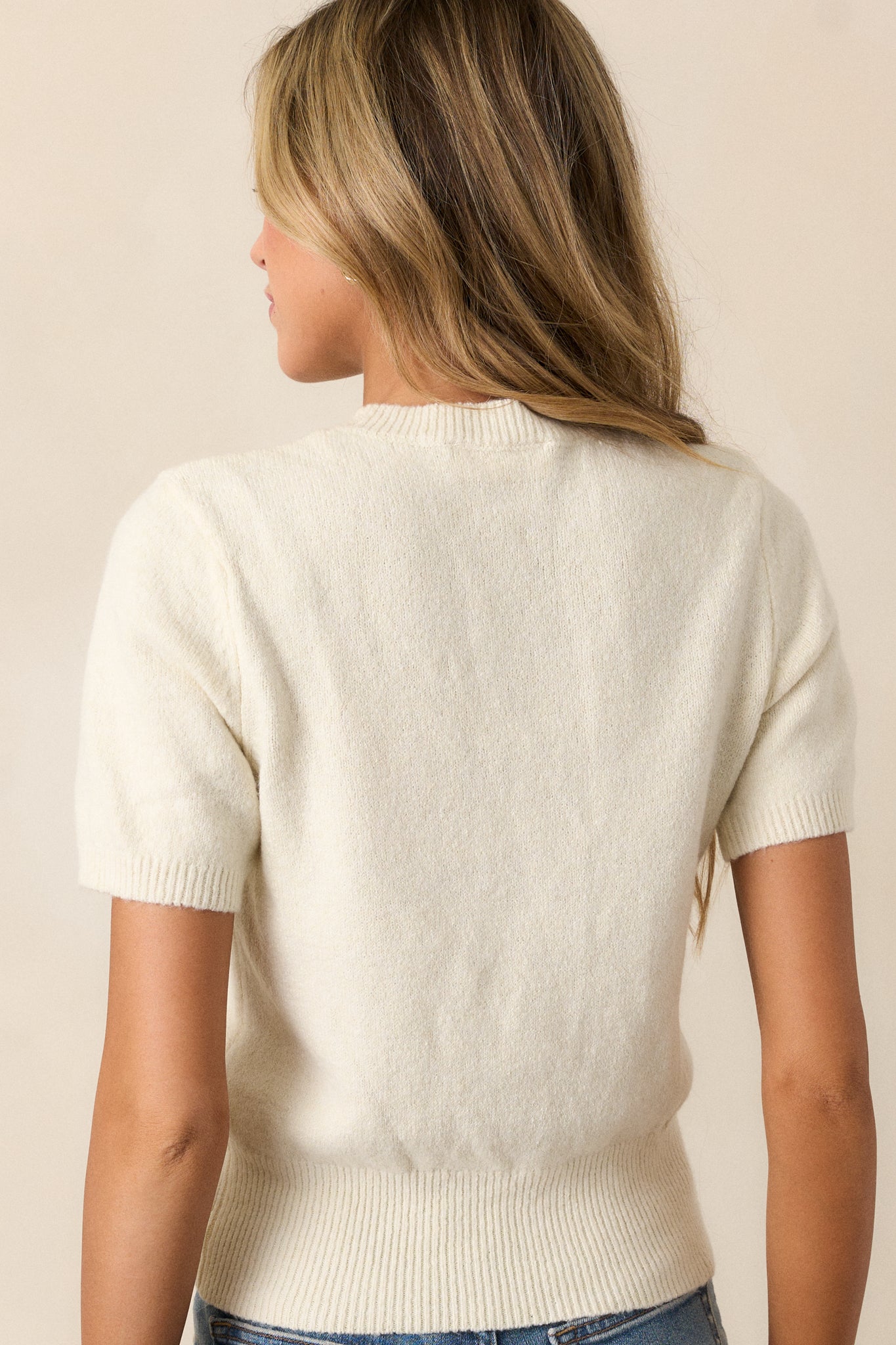 View of the back of the top, featuring the smooth ivory knit fabric and the continuous high neckline and ribbed hem details