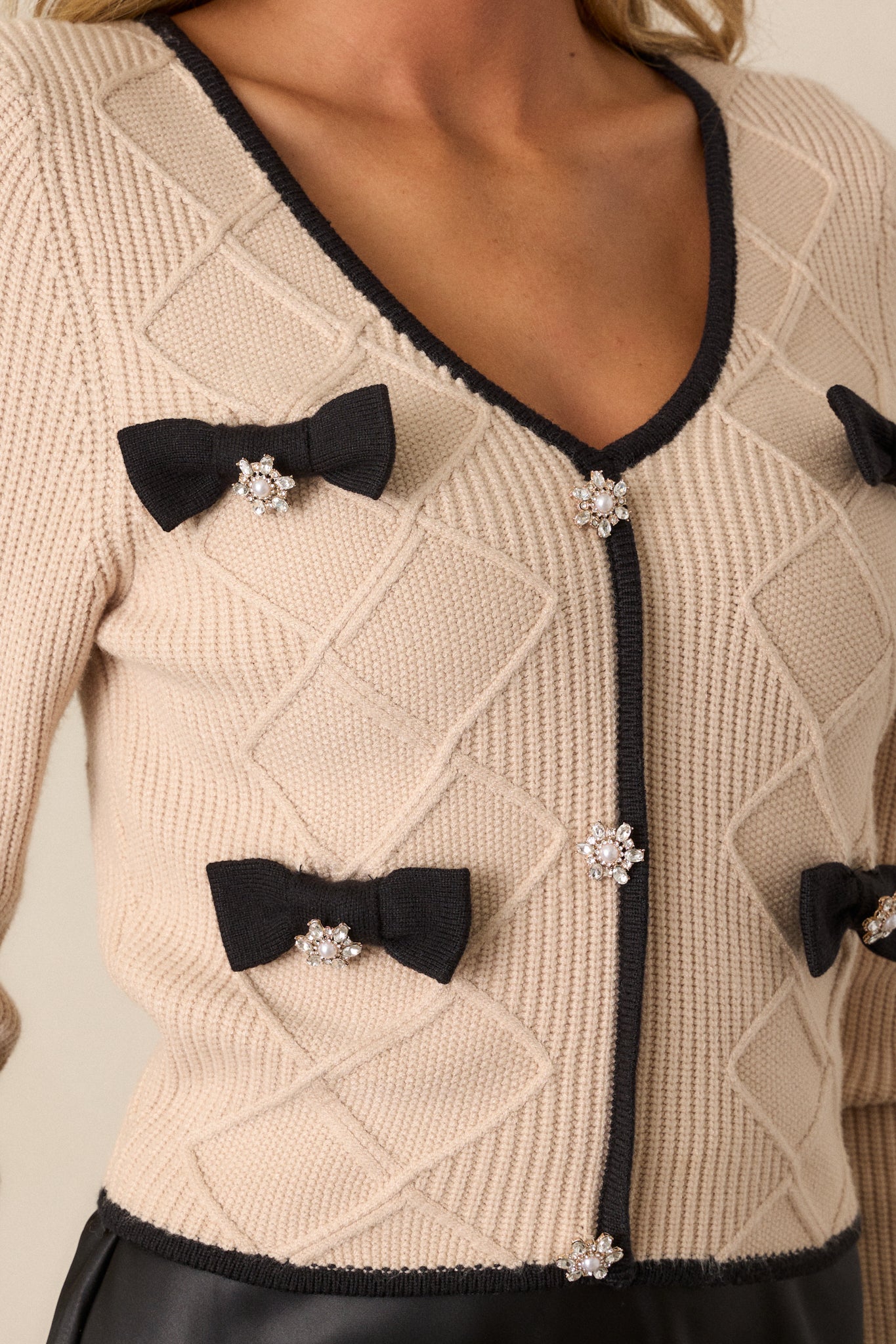 Close-up view of this beige sweater that features a v-neckline, a faux button front, black bow embellishments, faux pearl and rhinestone detailing, soft knit material, a black trim, long sleeves, and a cropped length.