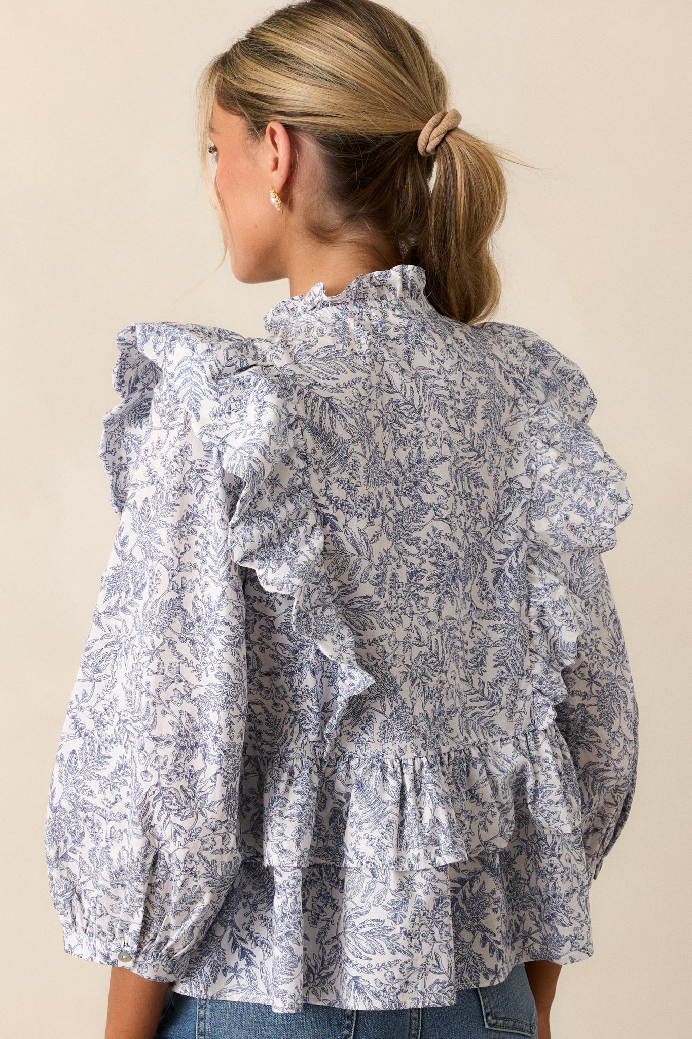 View of the back of the blouse, featuring the high ruffle neckline, ruffle shoulders, and the peplum fit that flares at the waist.