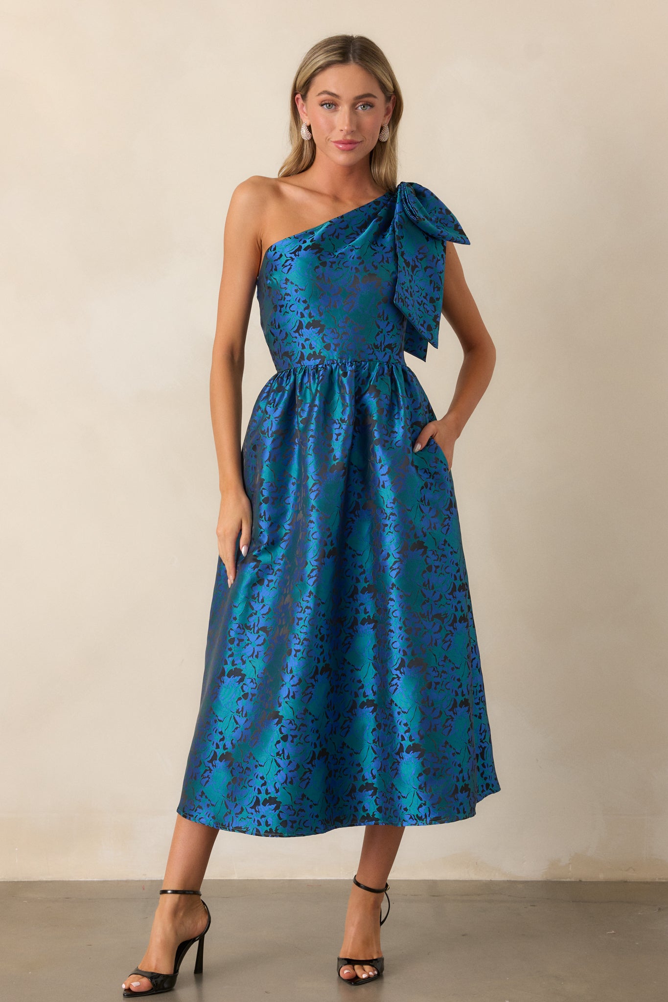 Full front view of the blue midi dress, featuring a one-shoulder neckline with a large bow embellishment, functional hip pockets, and a fit and flare silhouette. The jacquard fabric texture is visible in the light.