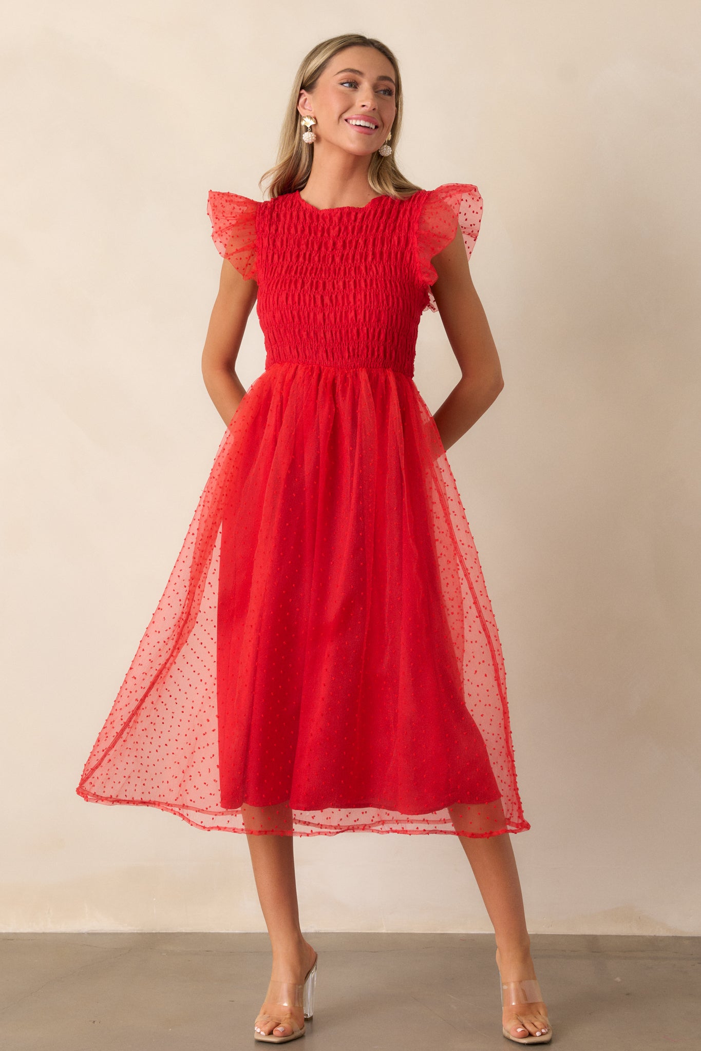 Front view of the red dress featuring a rounded neckline, a fully smocked bodice, and flutter sleeves, with a delicate polka dot tulle overlay adding texture to the design.