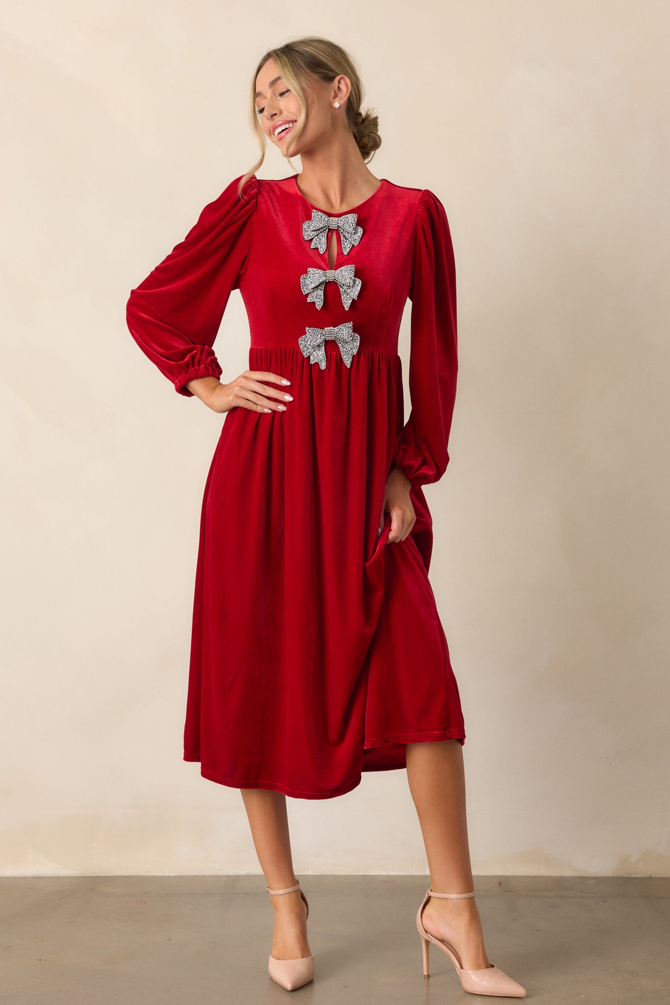 Full front view of the red velvet dress, showcasing the high rounded neckline, three rhinestone bows down the front bodice, and the relaxed skirt with a discrete back zipper.