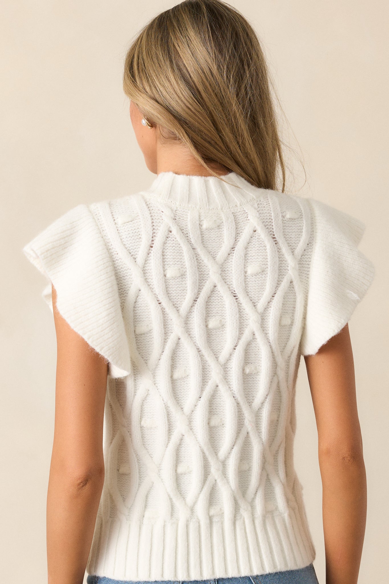 The back of the white sweater vest, highlighting the continuation of the cable knit design and ribbed hem detailing.