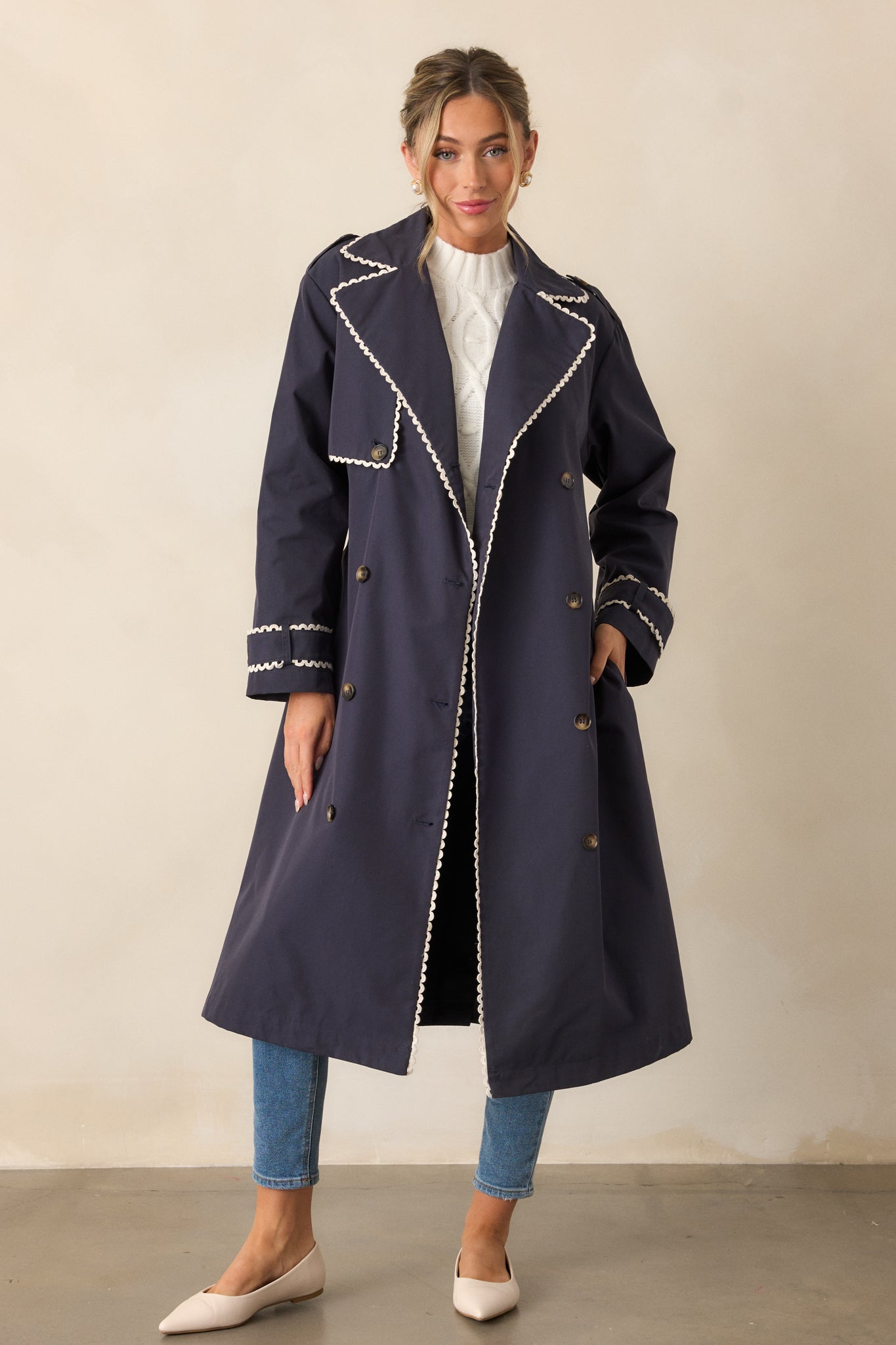 Navy trench coat shown in full body, highlighting its lapel collar and eight buttons down the front. The coat’s long sleeves are finished with button cuffs, and it has ricrac trim at the hem