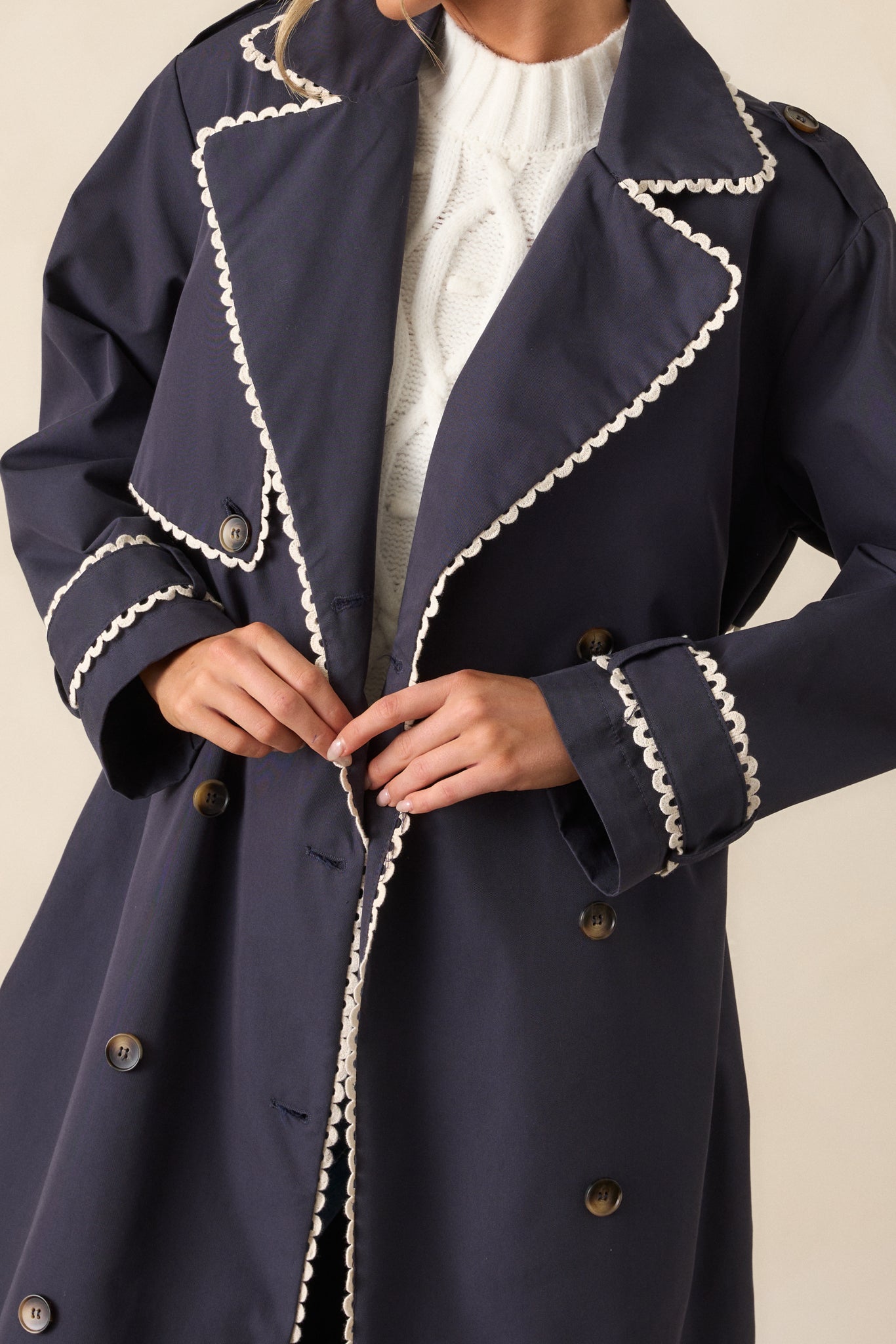 Close-up view of the navy trench coat’s lapel collar and button detailing. The image focuses on the coat's upper half, including the eight buttons and the elegant ricrac trim along the neckline.