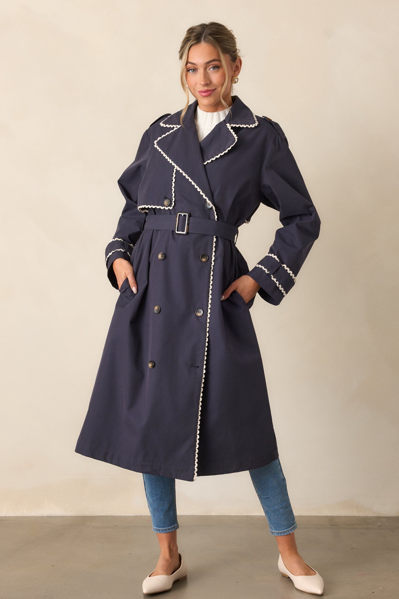 A full-body shot of a navy trench coat with a classic lapel collar, button front closure with eight buttons, and a self-tie waistband. The long sleeves have button cuffs and ricrac trim is visible along the edges.