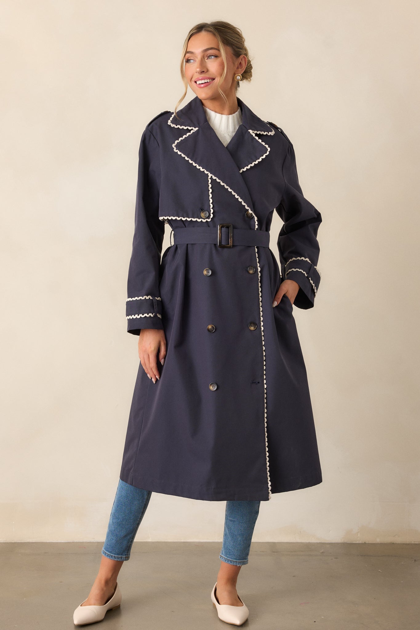 A full-body view of a navy trench coat with a notched lapel collar and double-breasted button front. The coat is styled with an optional self-tie belt and features ivory ricrac trim along the edges. The long sleeves have button cuffs, completing the sleek, sophisticated look.