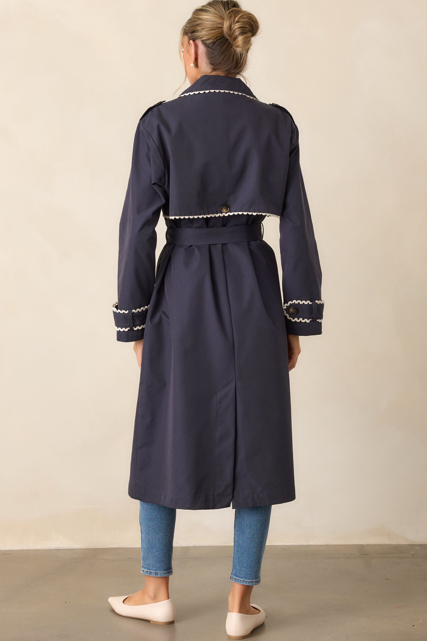 Back view of a navy trench coat with a self-tie waistband, showcasing the eight button front closure. The long sleeves with button cuffs and the ricrac trim along the edges are also visible from behind.