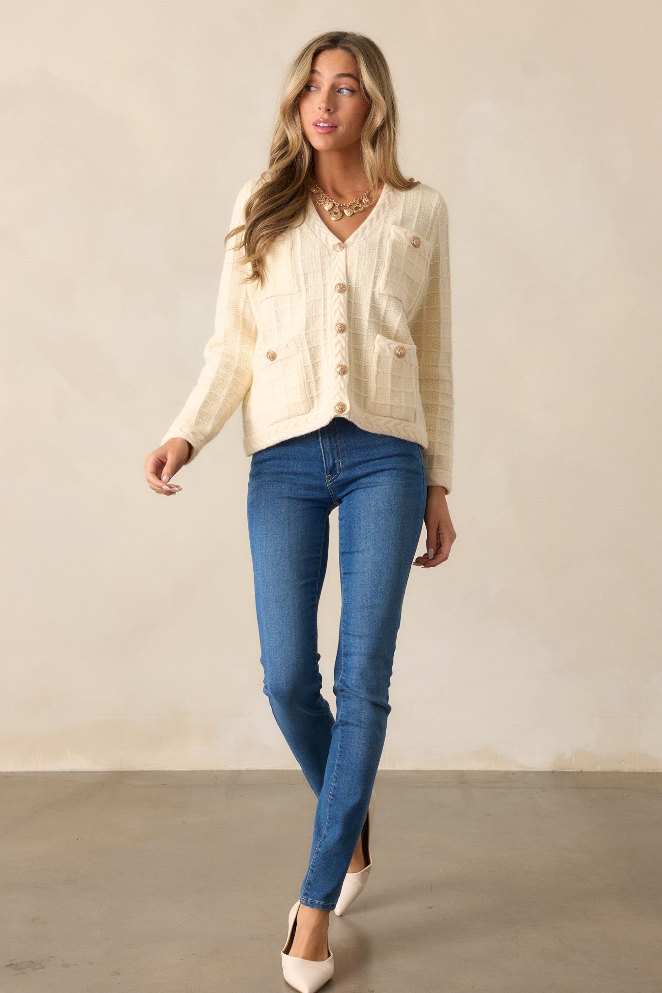 A full view of the ivory cardigan featuring a v-neckline, functional gold button front, and long sleeves. The cable knit grid design and relaxed silhouette are visible.